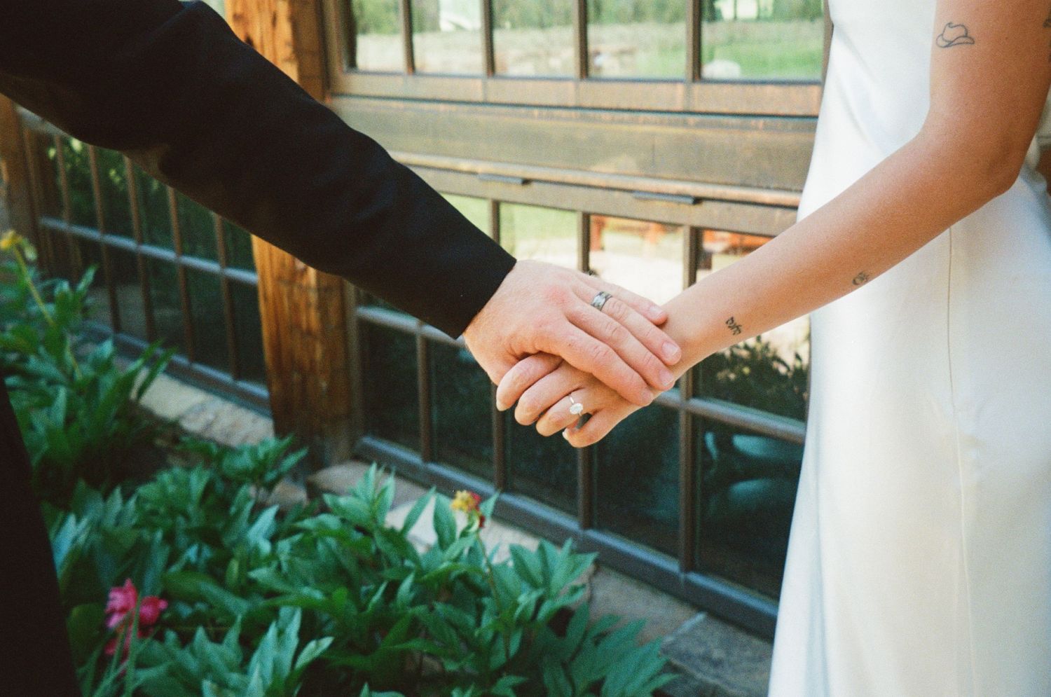 Close-up of bride and groom holding hands showing wedding ring with greenery in background.