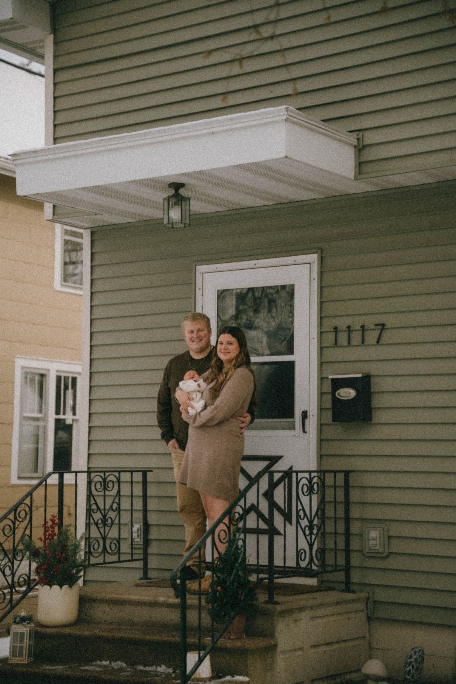 Happy couple holding newborn baby on front porch of green house numbered 1117.