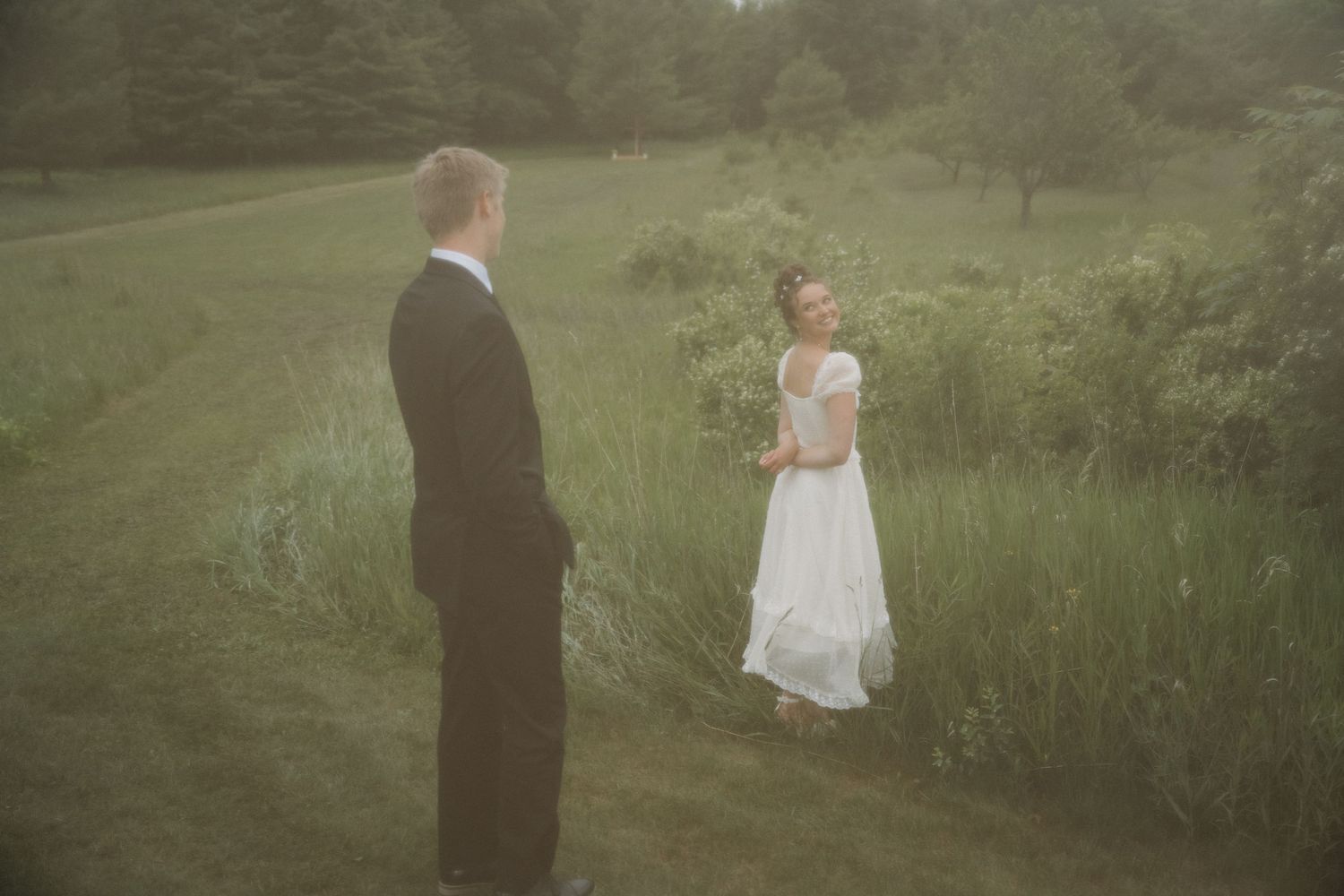 A bride in a white gown and groom in a black suit stand facing each other in a misty green outdoor setting.