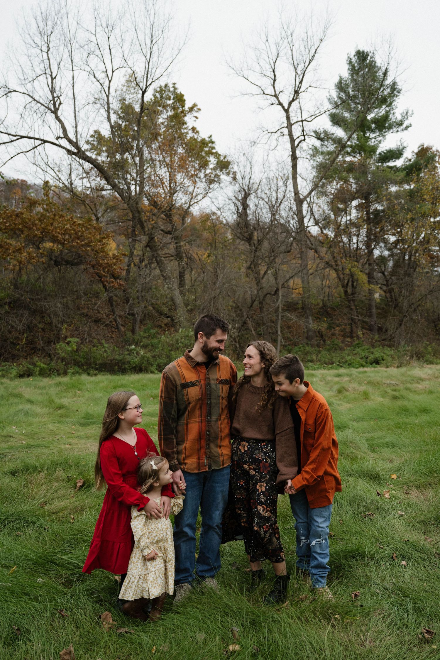 Happy family of five stands together in a green autumn field surrounded by bare trees on an overcast day.