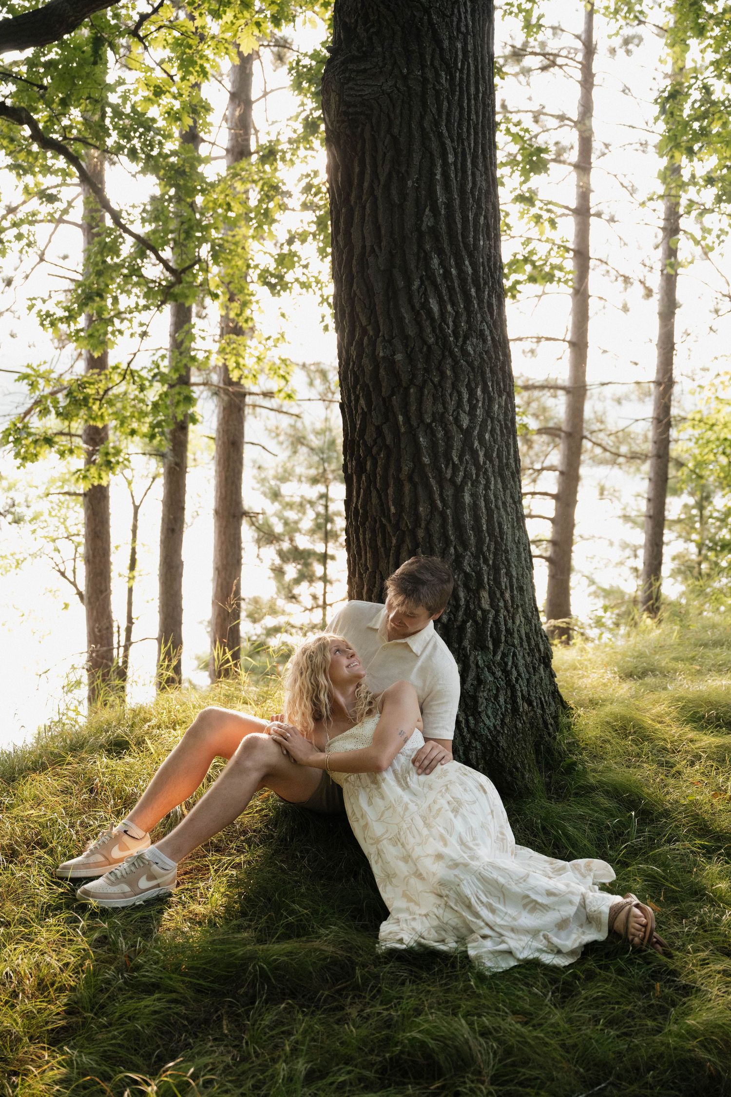 Couple sitting together against large tree trunk in sunlit forest, woman in white floral dress.