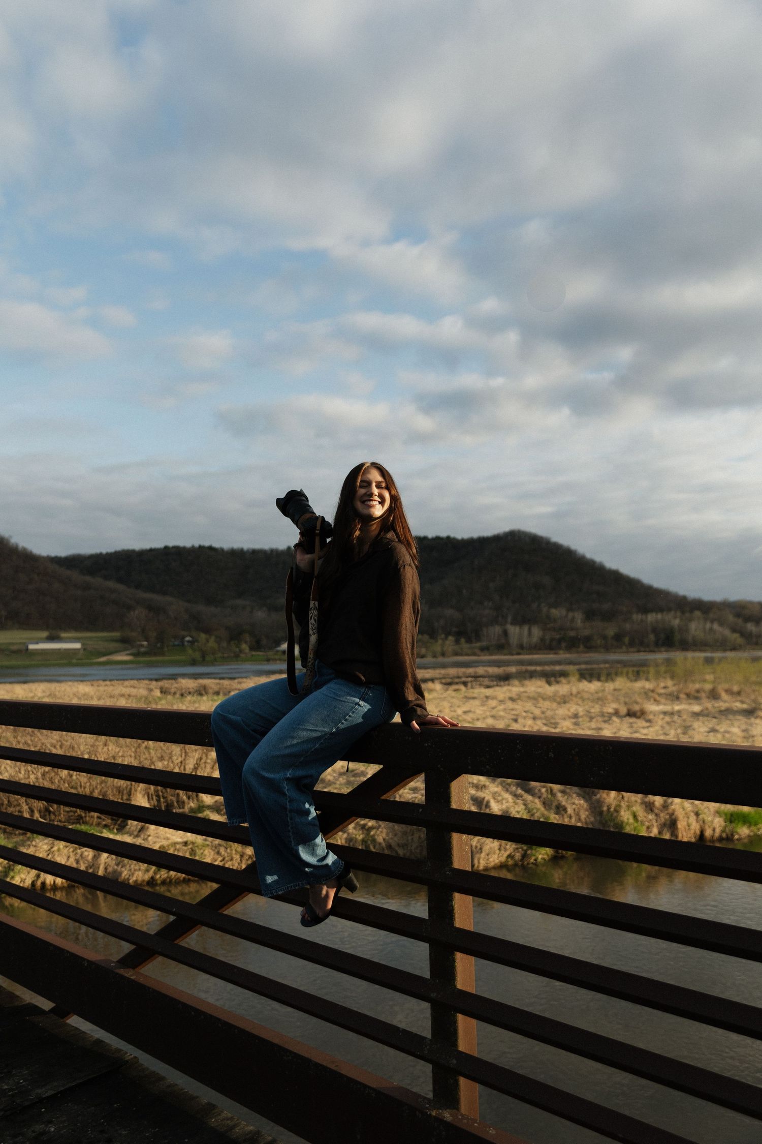 Smiling woman with camera sitting on fence with mountain landscape behind her.