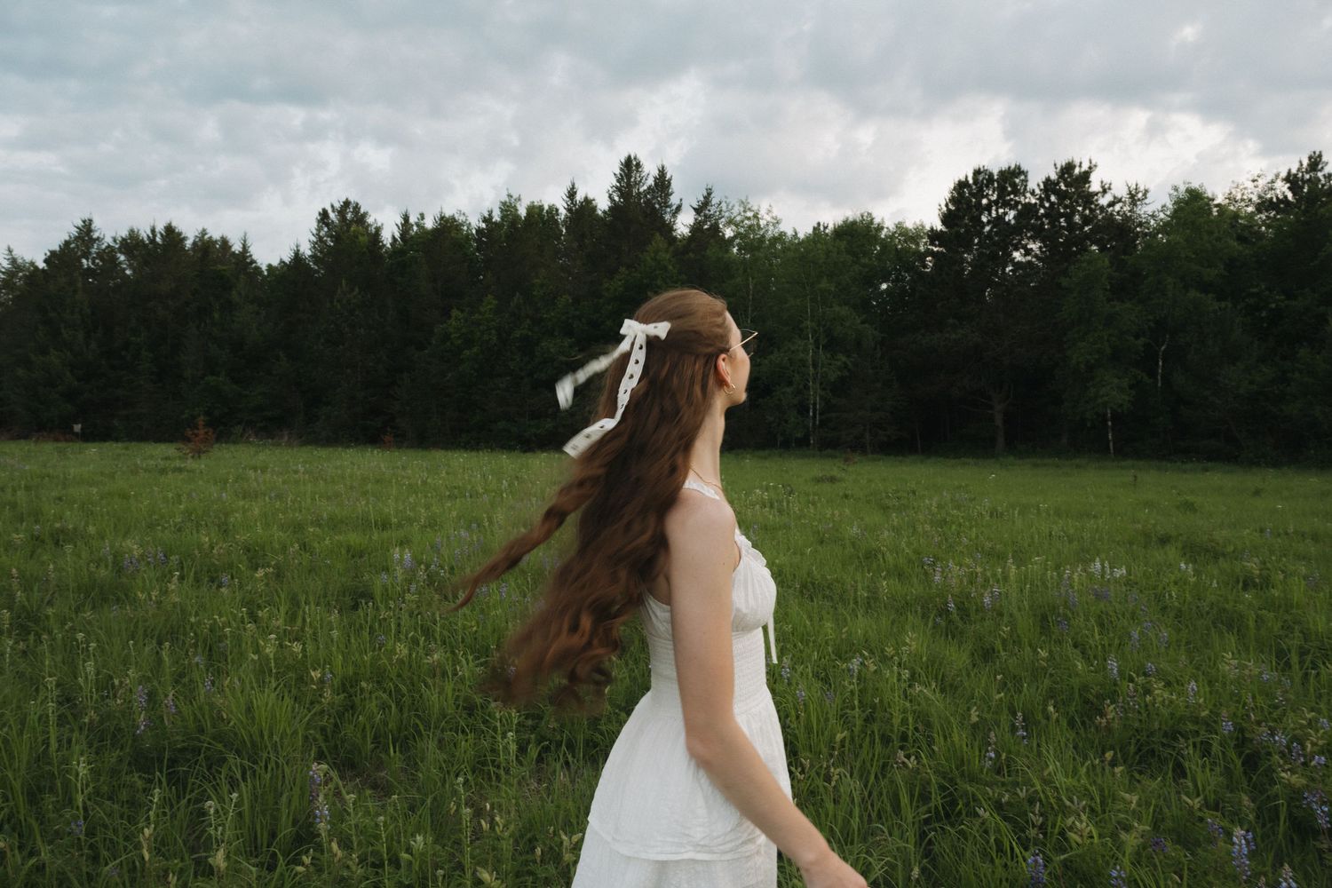 Young woman in white dress with ribbon hair accessory walks through a green field with trees in the background.