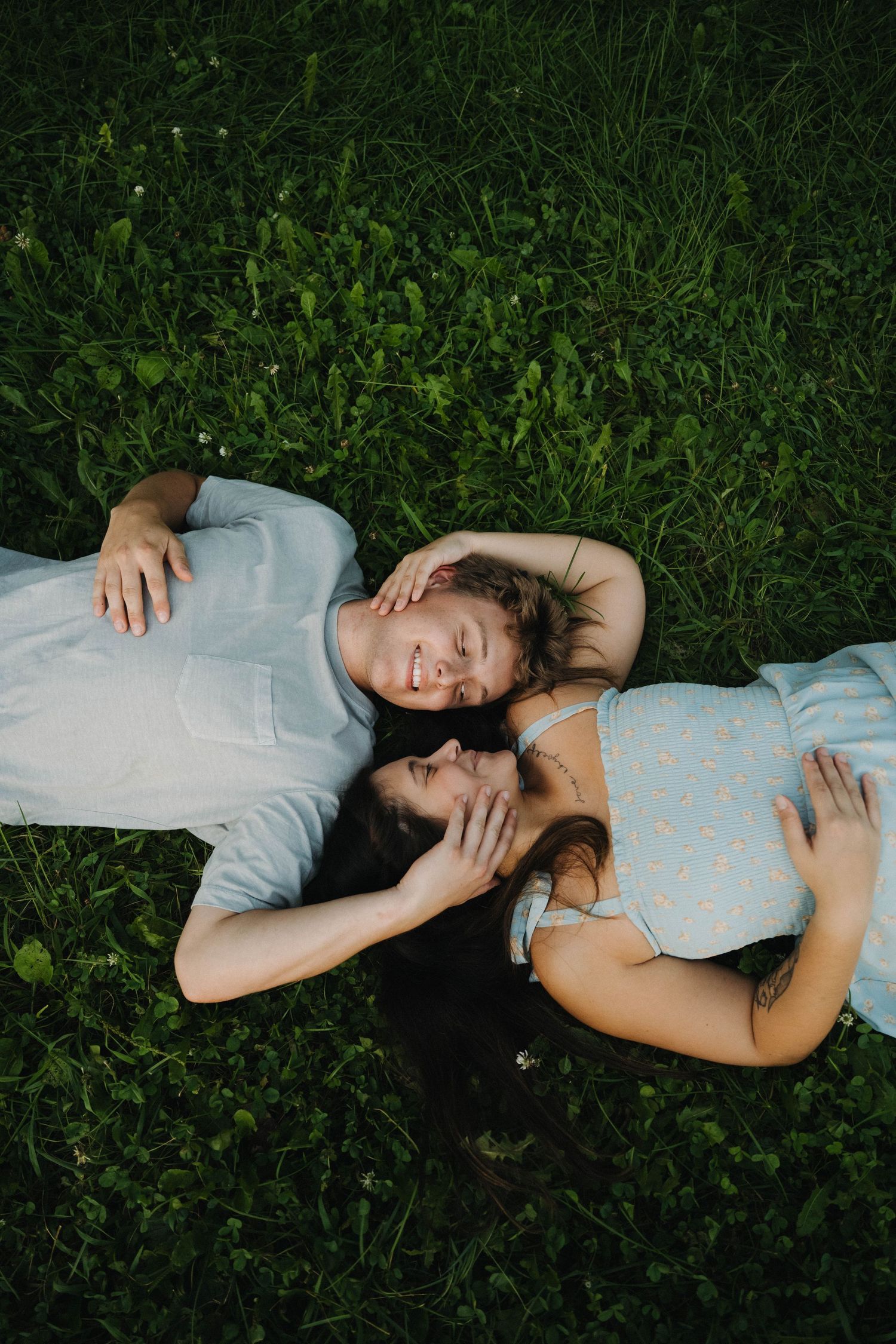 A couple lying on green grass, smiling and touching heads together in a romantic outdoor moment.