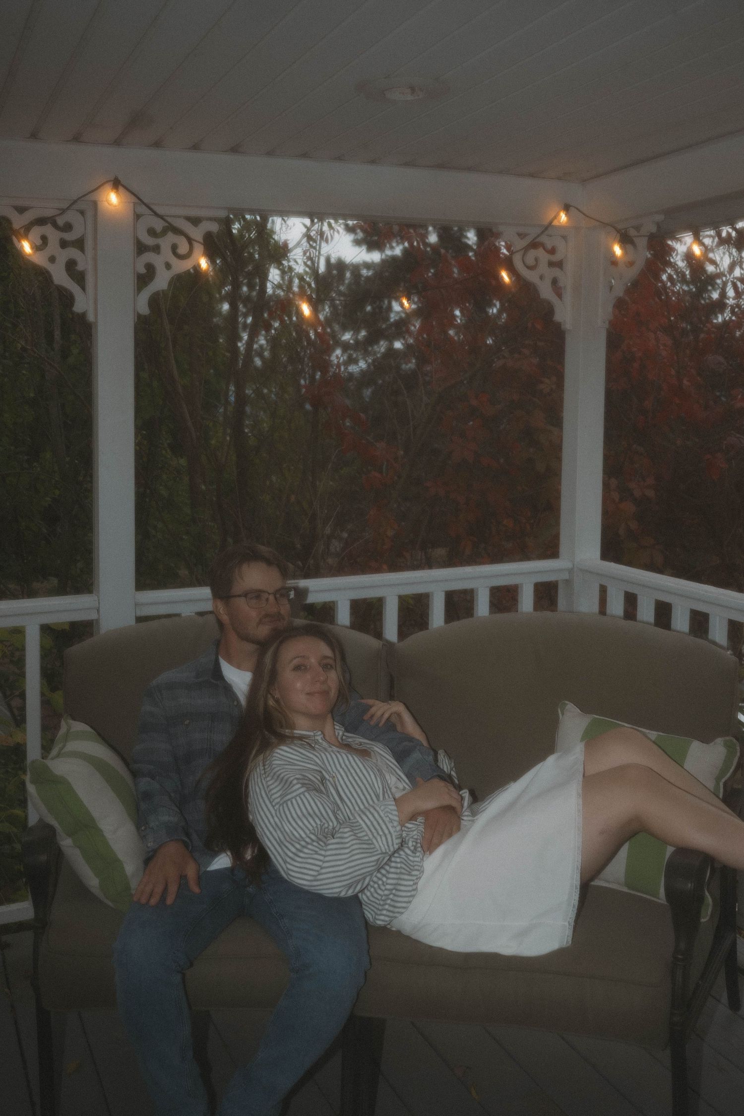 A couple relaxing together on a porch swing surrounded by string lights and autumn foliage at dusk.