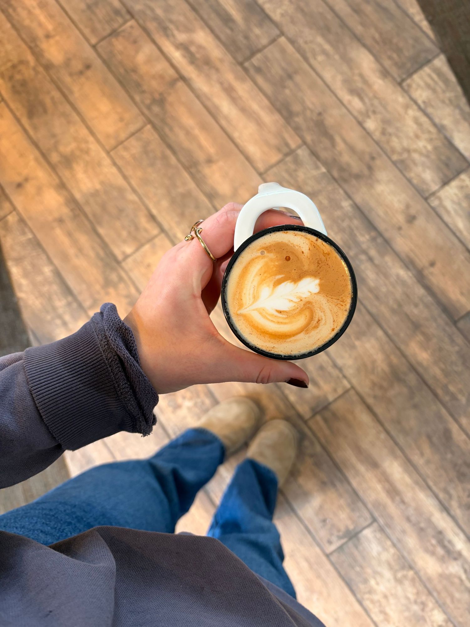 Hand holding a latte art coffee cup above a wooden floor with blue jeans visible.