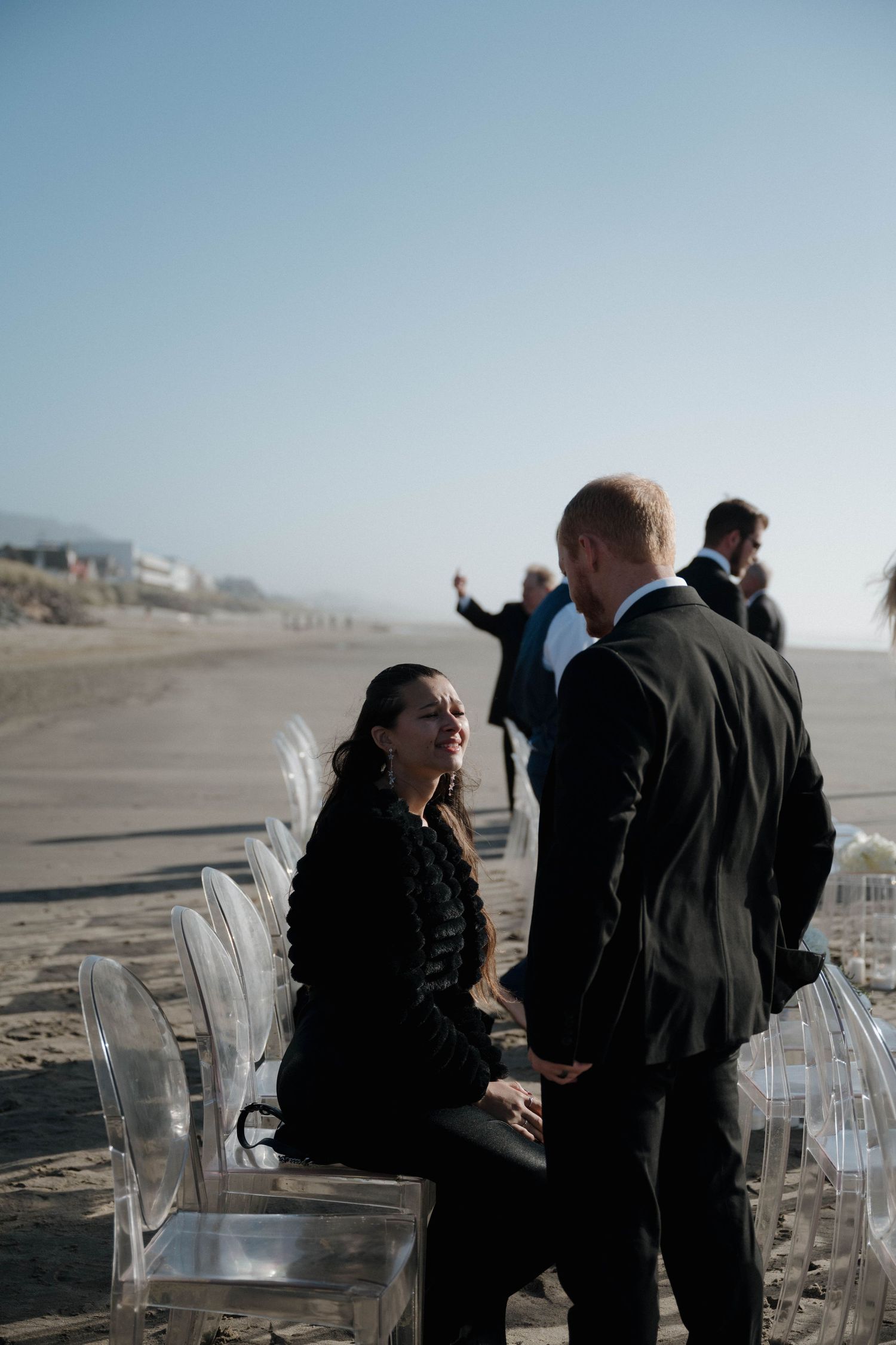 A woman in black sits on a clear chair at a beach wedding ceremony while a man in a suit stands nearby.