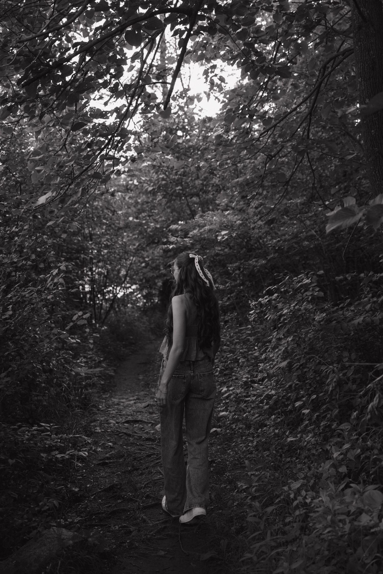 Black and white photo of a young woman walking barefoot along a dark, tree-lined forest path.