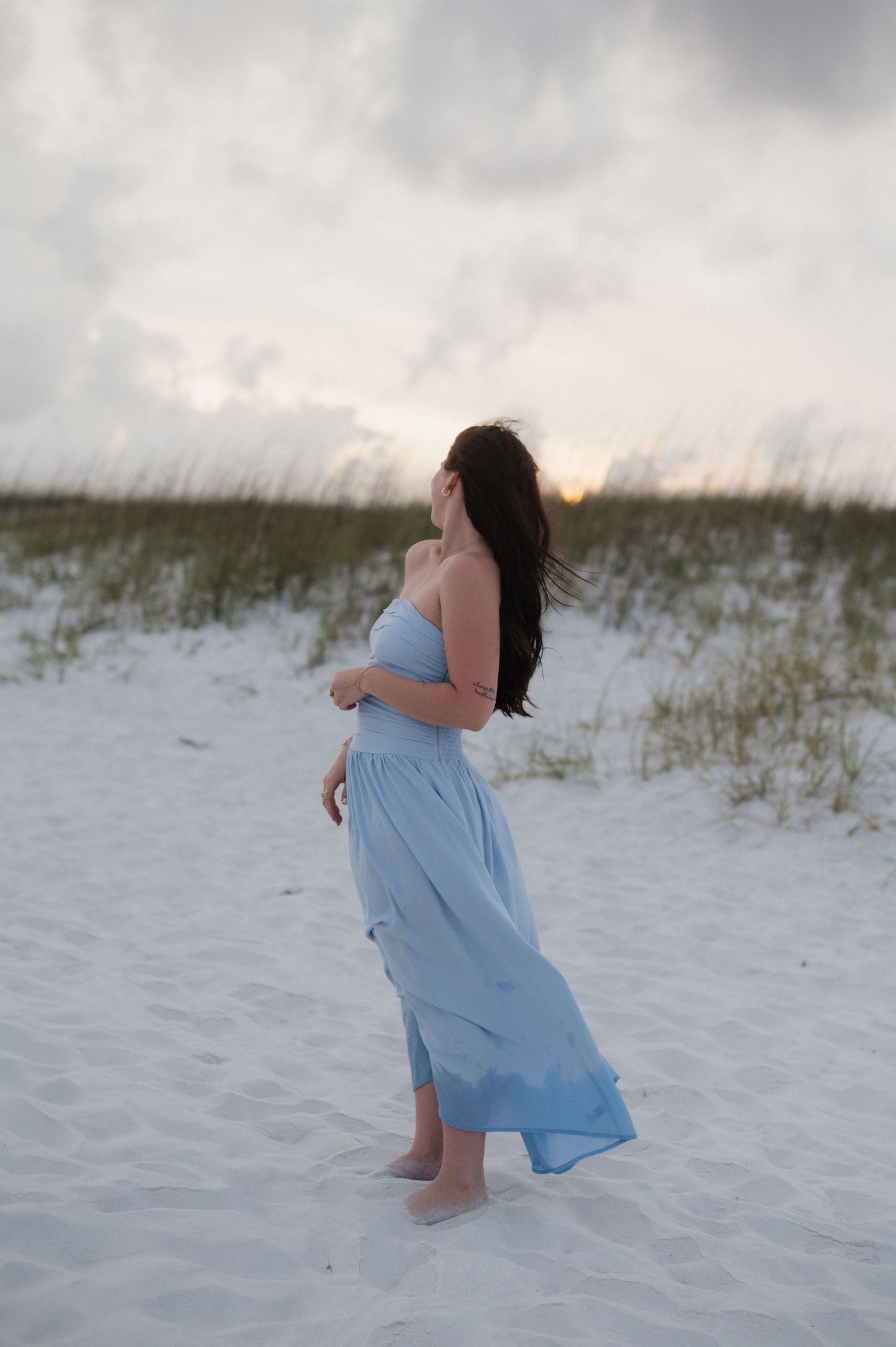 Woman in flowing blue strapless maxi dress standing barefoot on white sand beach at sunset.