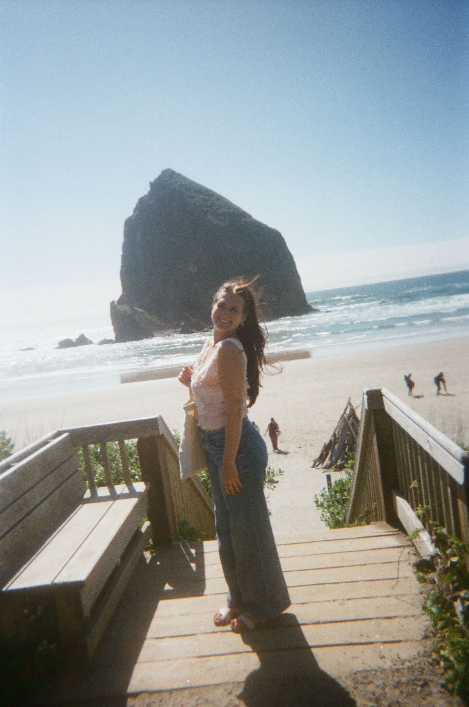 Smiling brunette woman in jeans on beach boardwalk stairs with iconic Haystack Rock at Cannon Beach.