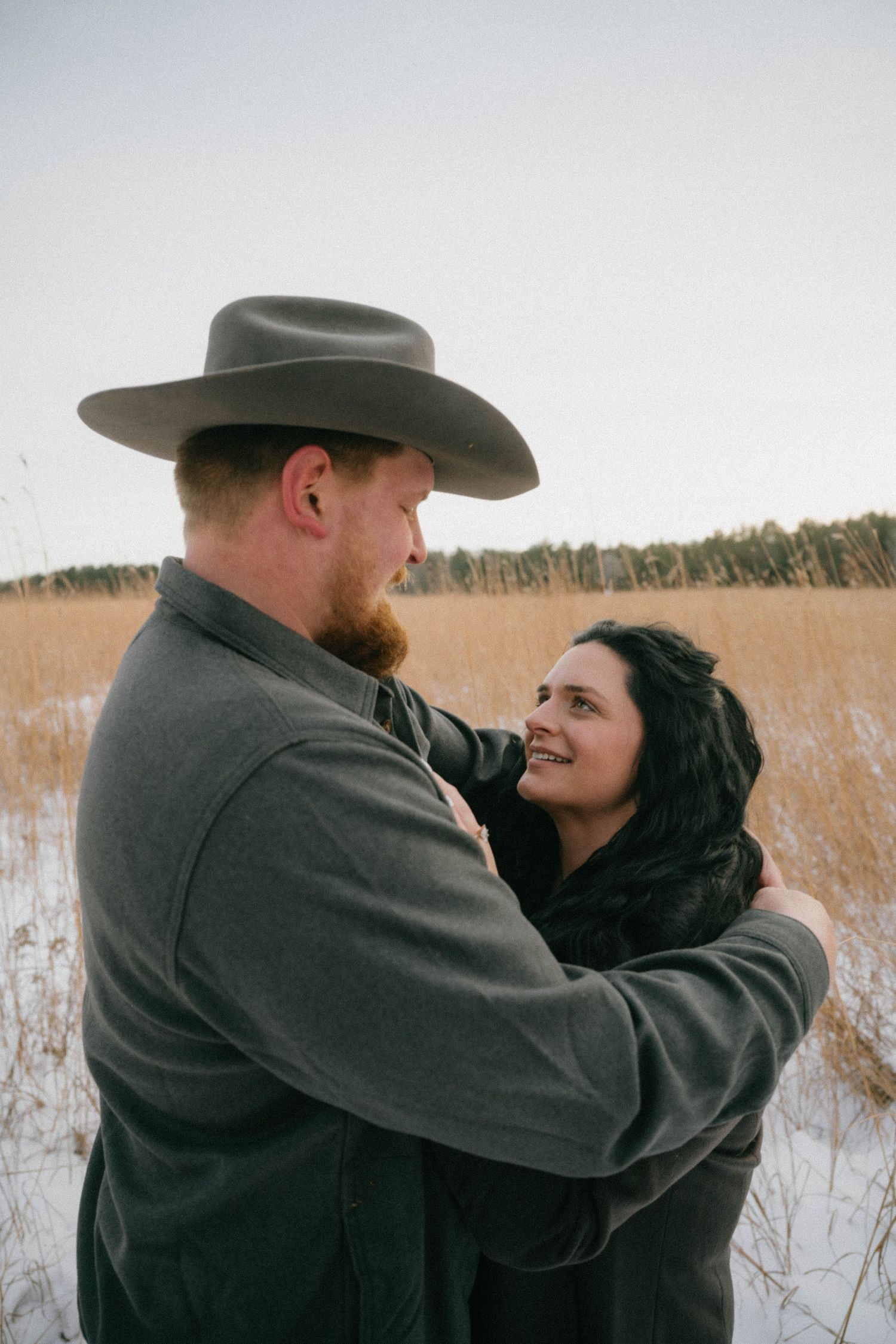 Couple embracing in snowy winter field, man in cowboy hat and gray jacket, woman smiling up at him.