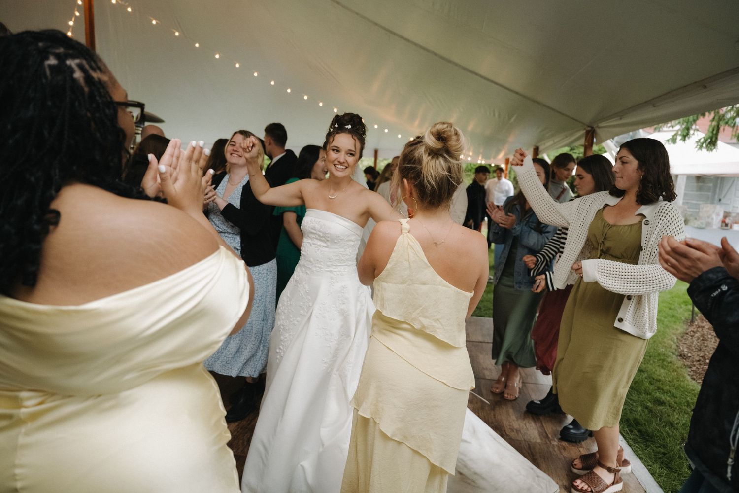 Bride dances joyfully with guests under lit tent at wedding reception.