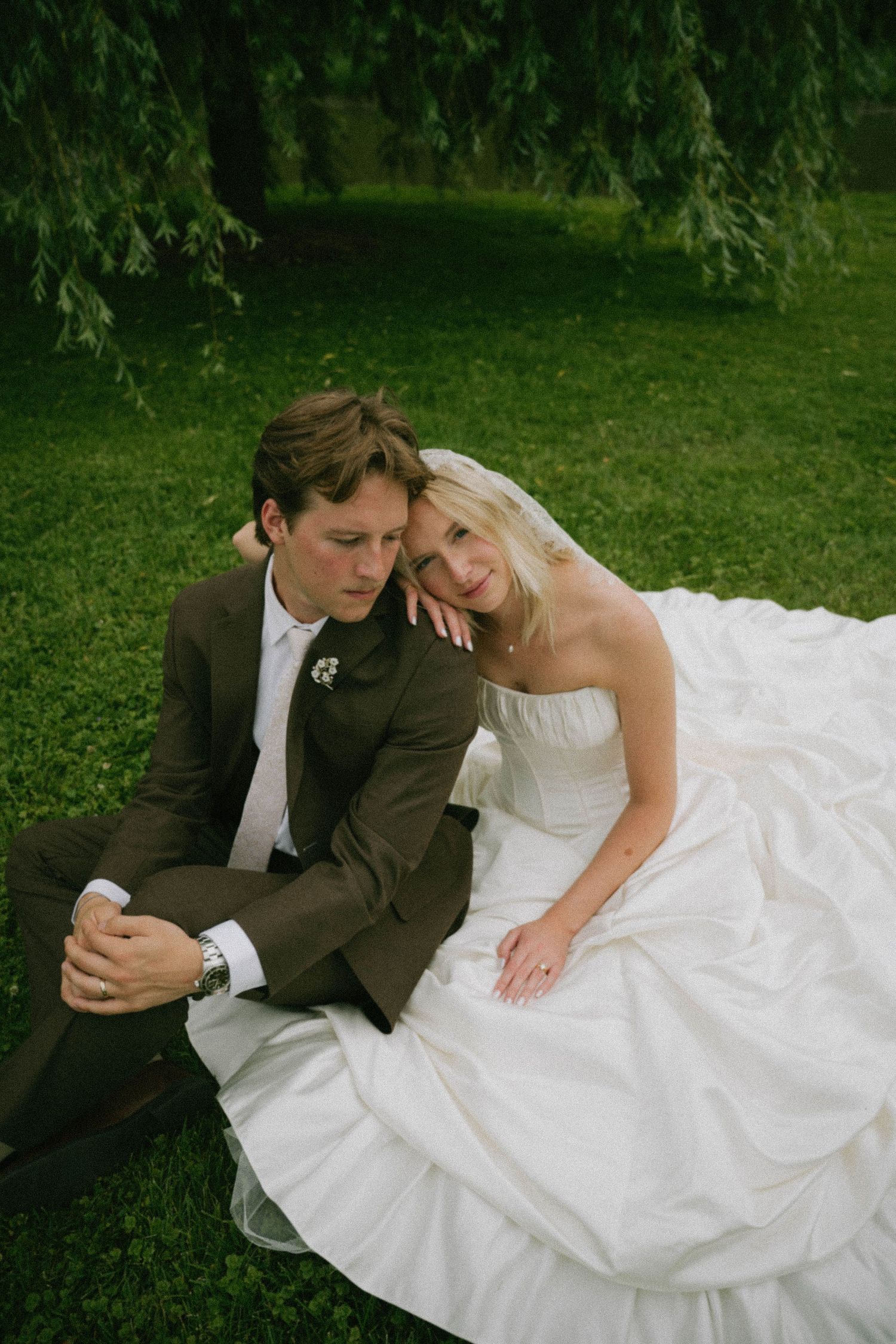 Bride and groom sitting together on green grass under a willow tree, bride in white ball gown, groom in brown suit.