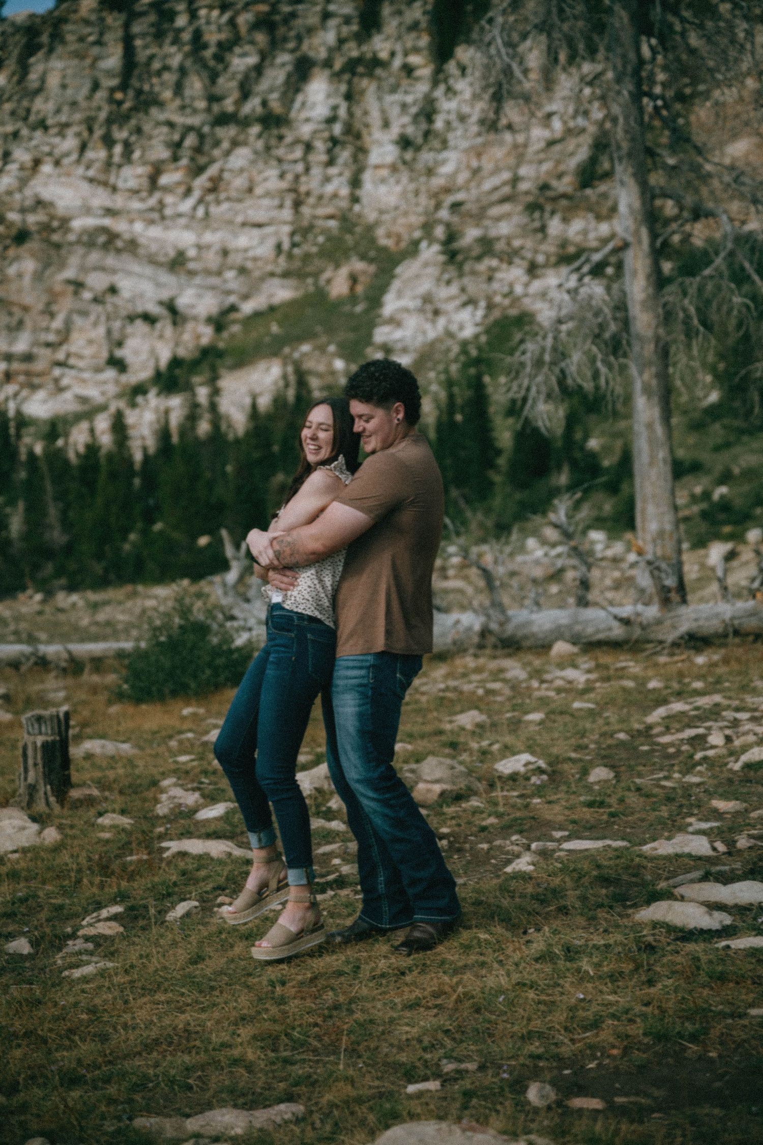 A couple embraces and laughs together in a rocky mountain meadow with pine trees in the background.