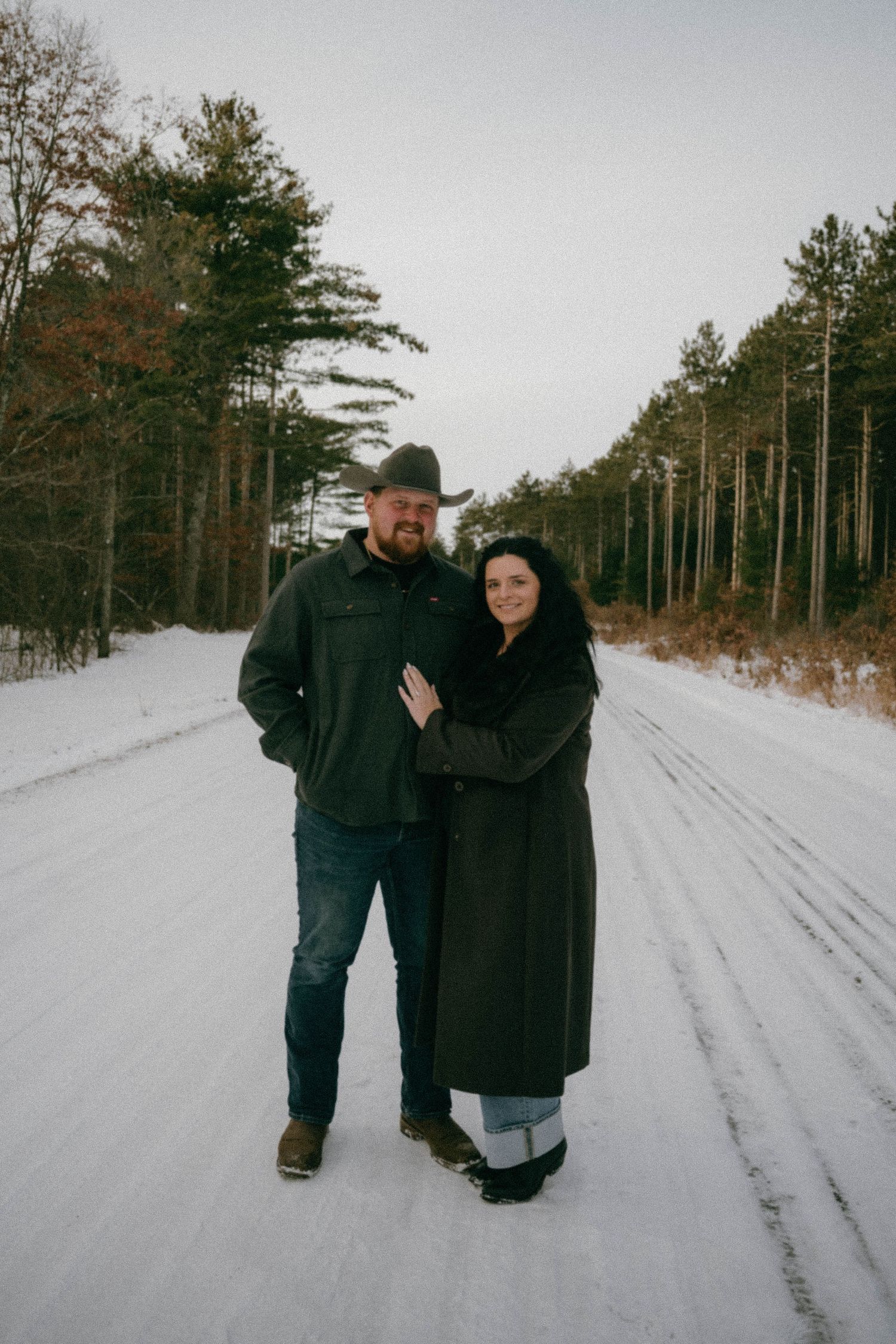 Couple posing together on a snowy forest road in winter coats.