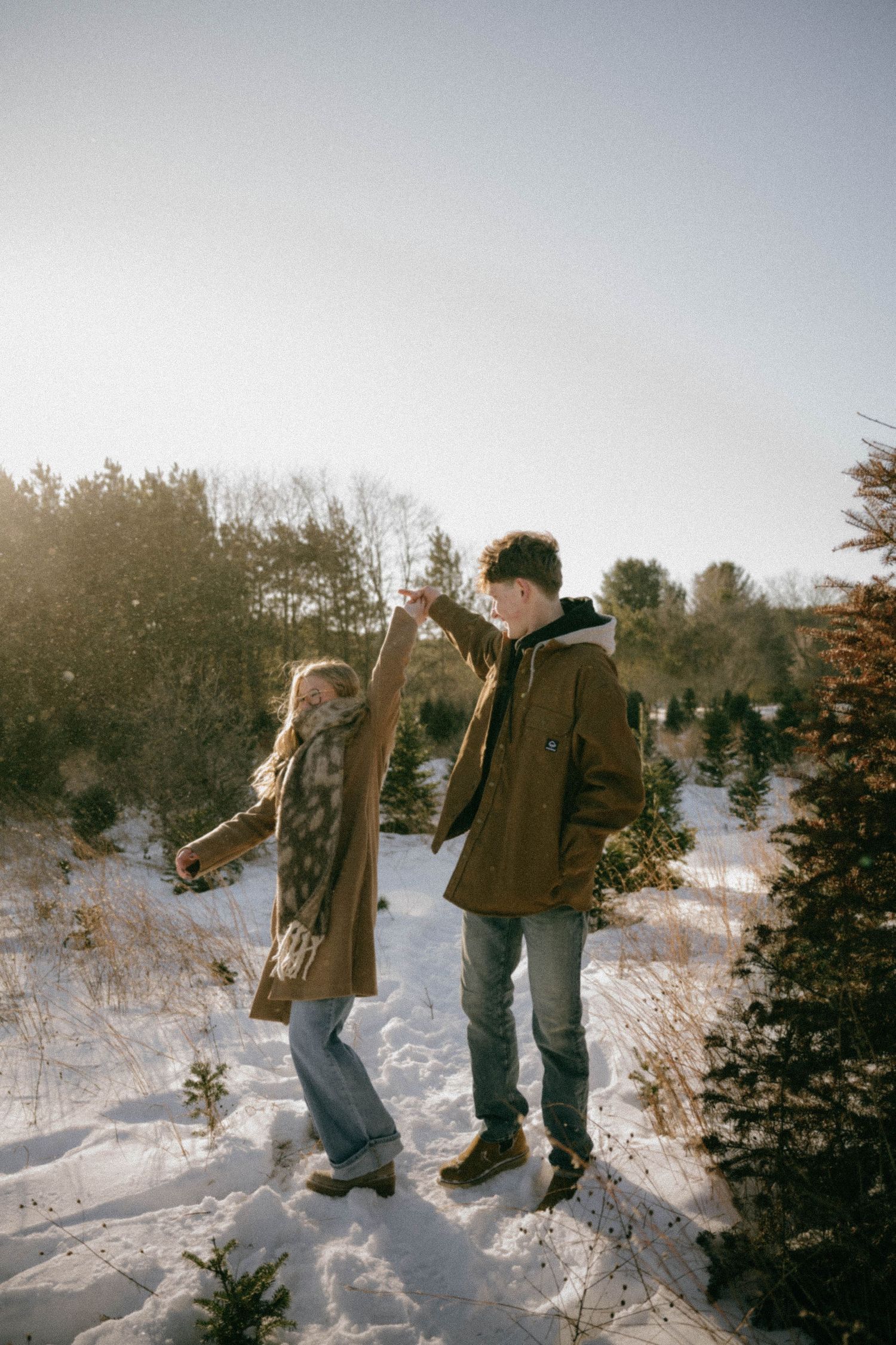 Couple dances joyfully in a snowy winter landscape surrounded by pine trees in golden sunlight.