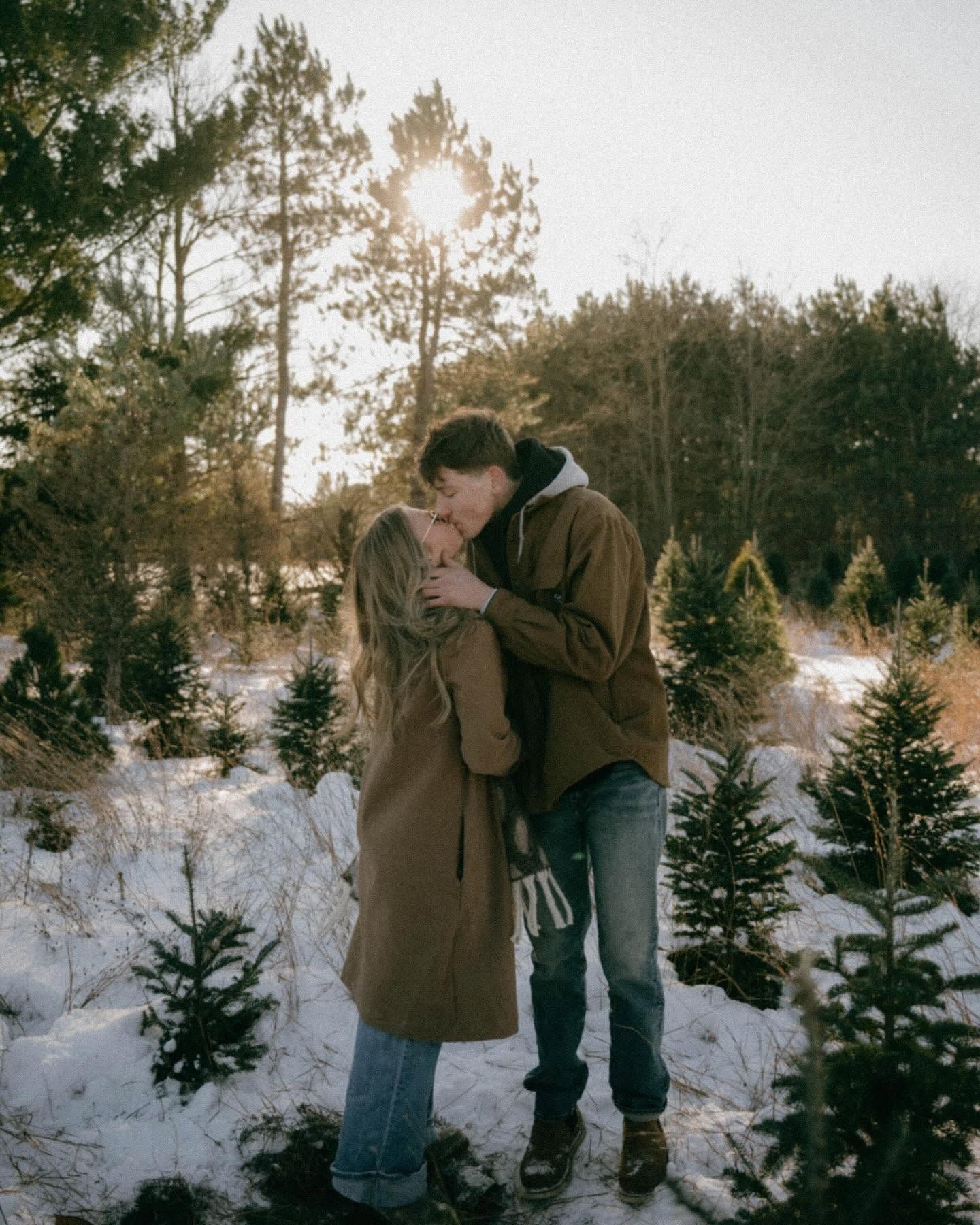 Couple kissing in snowy Christmas tree farm at golden hour, wearing cozy winter coats amid evergreen trees.