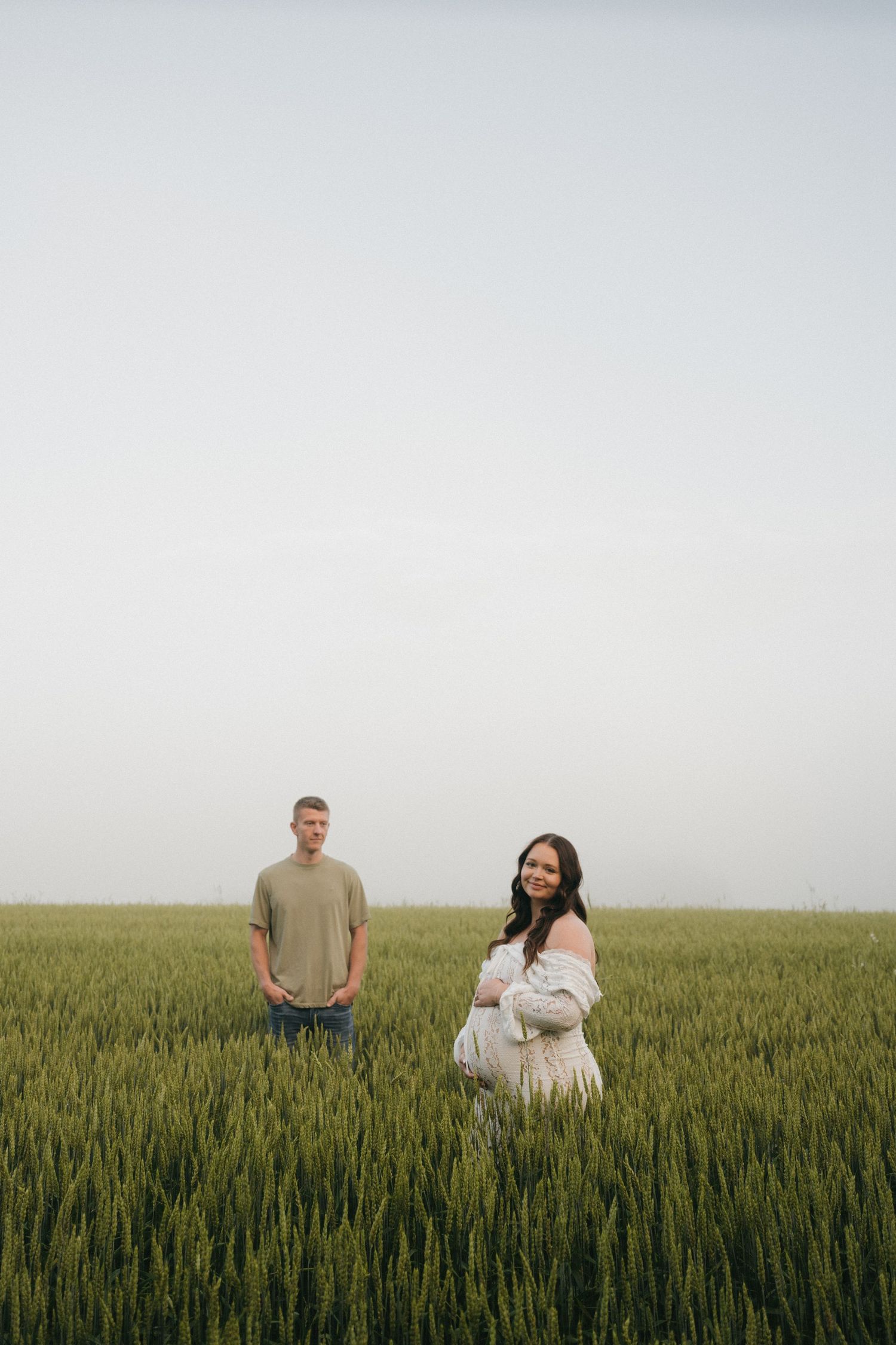 Pregnant woman in white dress smiles in green wheat field while man stands behind her on a foggy day.