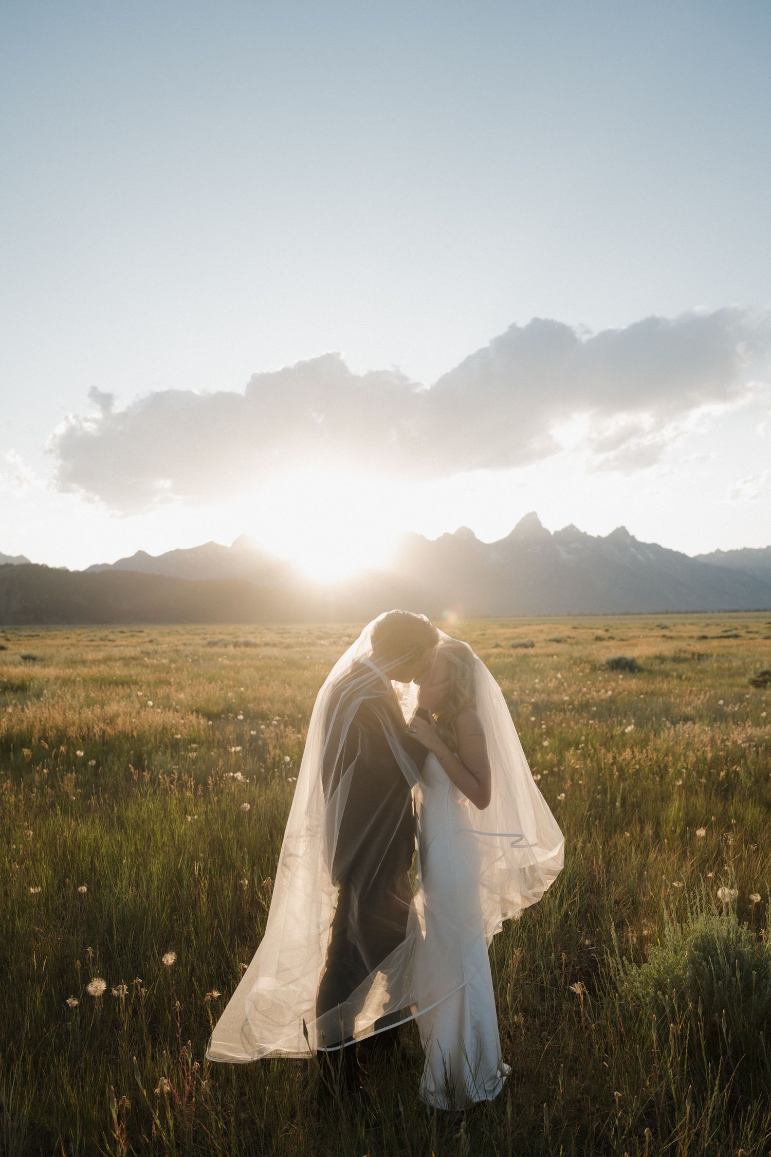 Couple kisses under flowing veil in scenic mountain meadow with dramatic sunset backdrop.