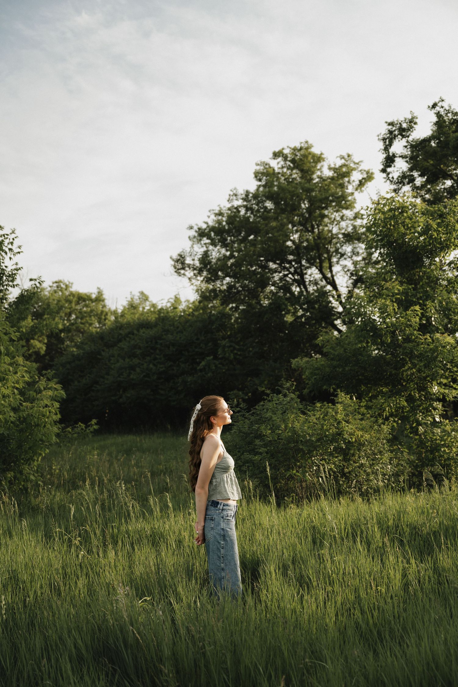 Woman in casual tank top and denim skirt standing in tall green grass field at golden hour.