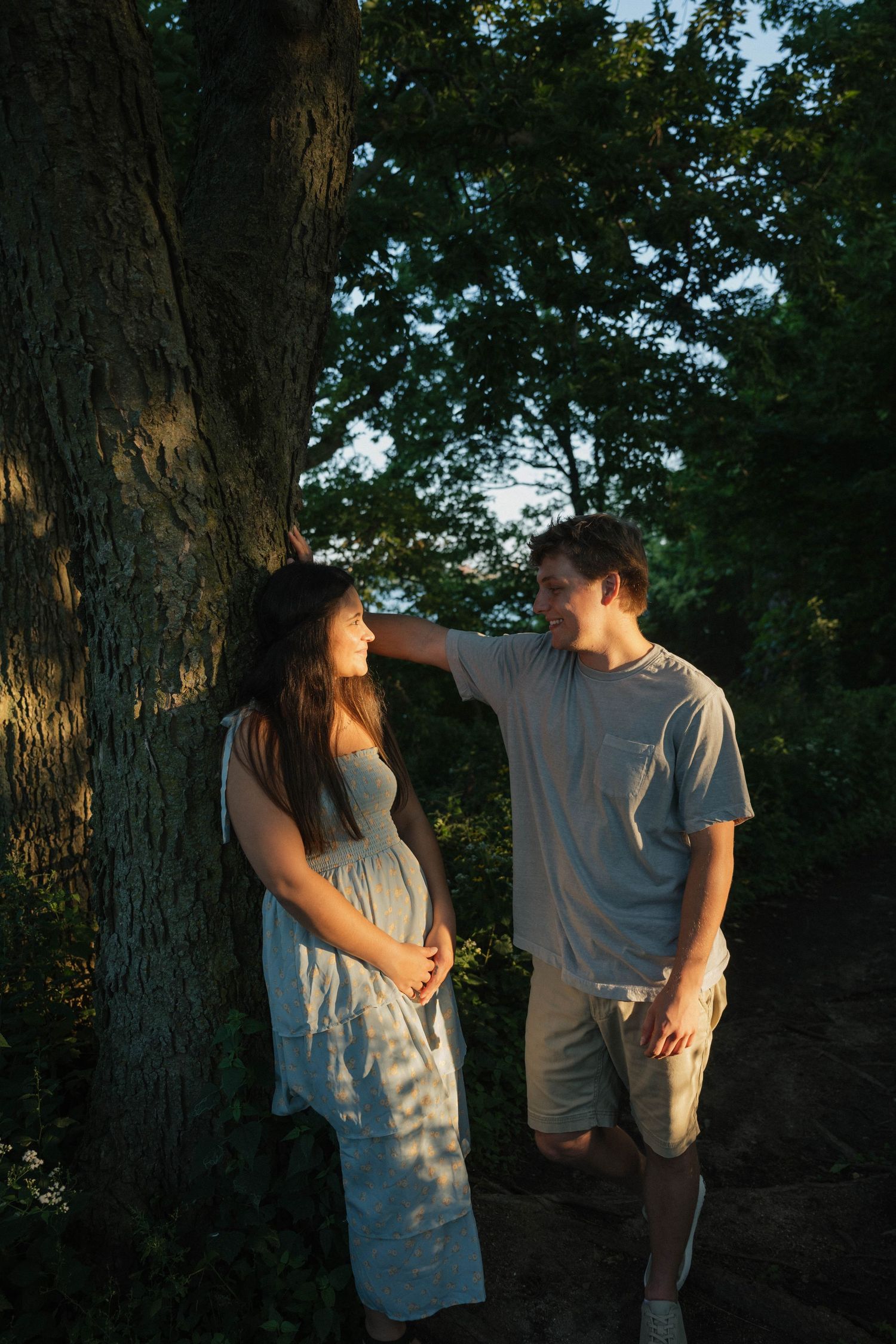 Couple sharing a tender moment leaning against large tree trunk in golden evening sunlight.