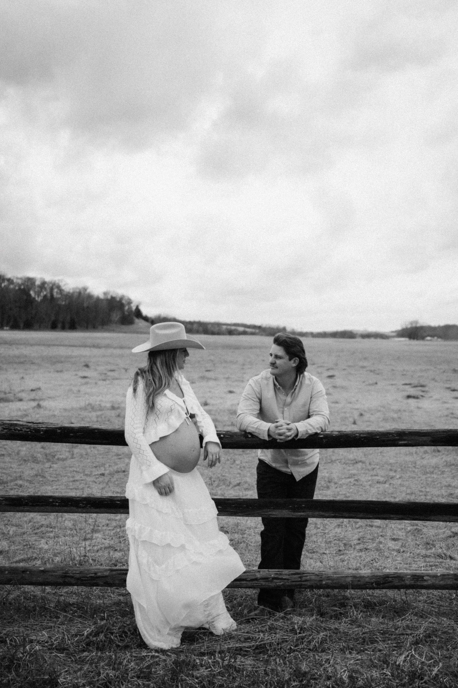 Black and white maternity photo of couple at rustic fence in open field under cloudy sky.