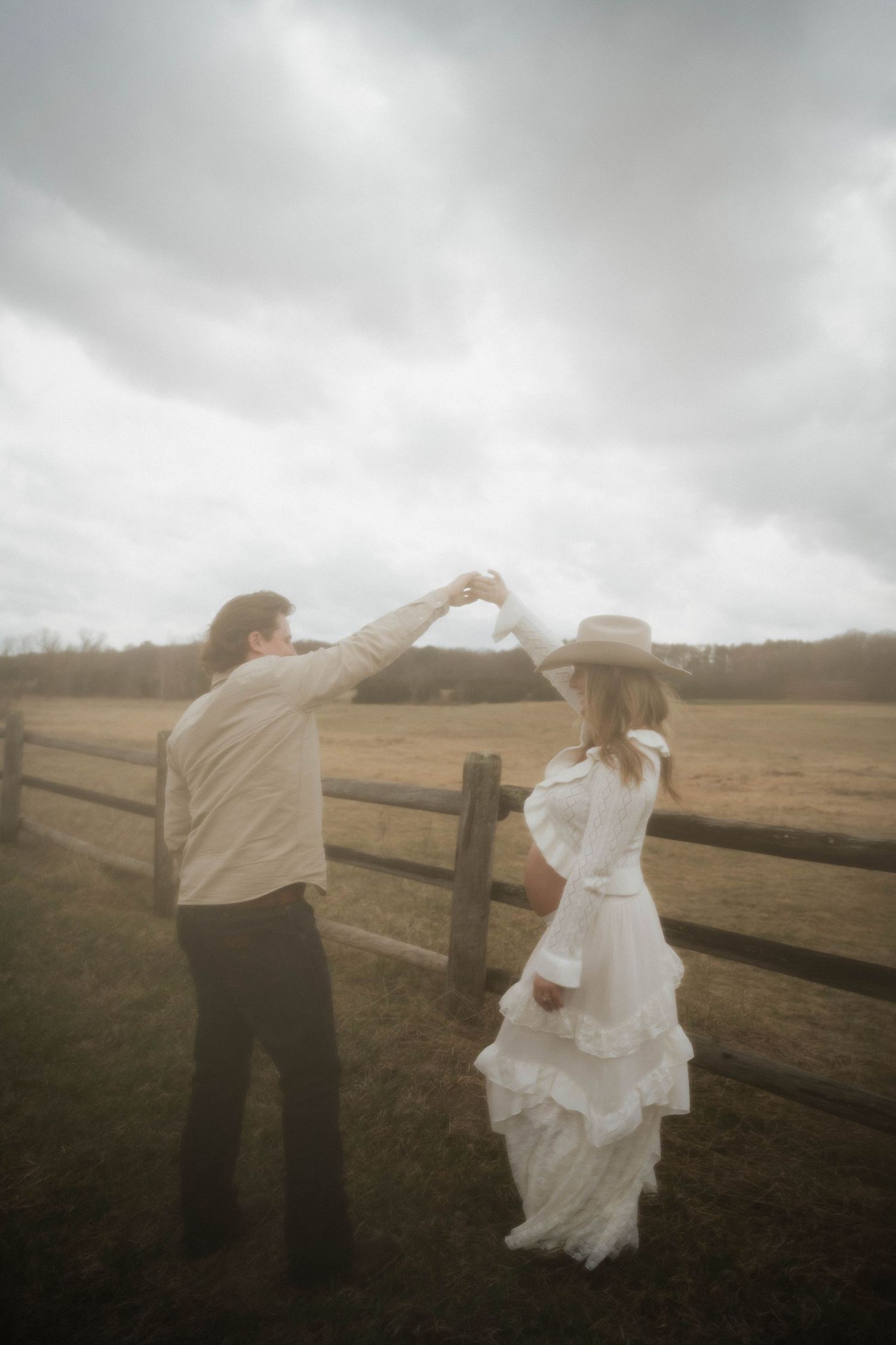 Couple dancing by a rustic wooden fence in an open field, woman in white dress and cowboy hat.