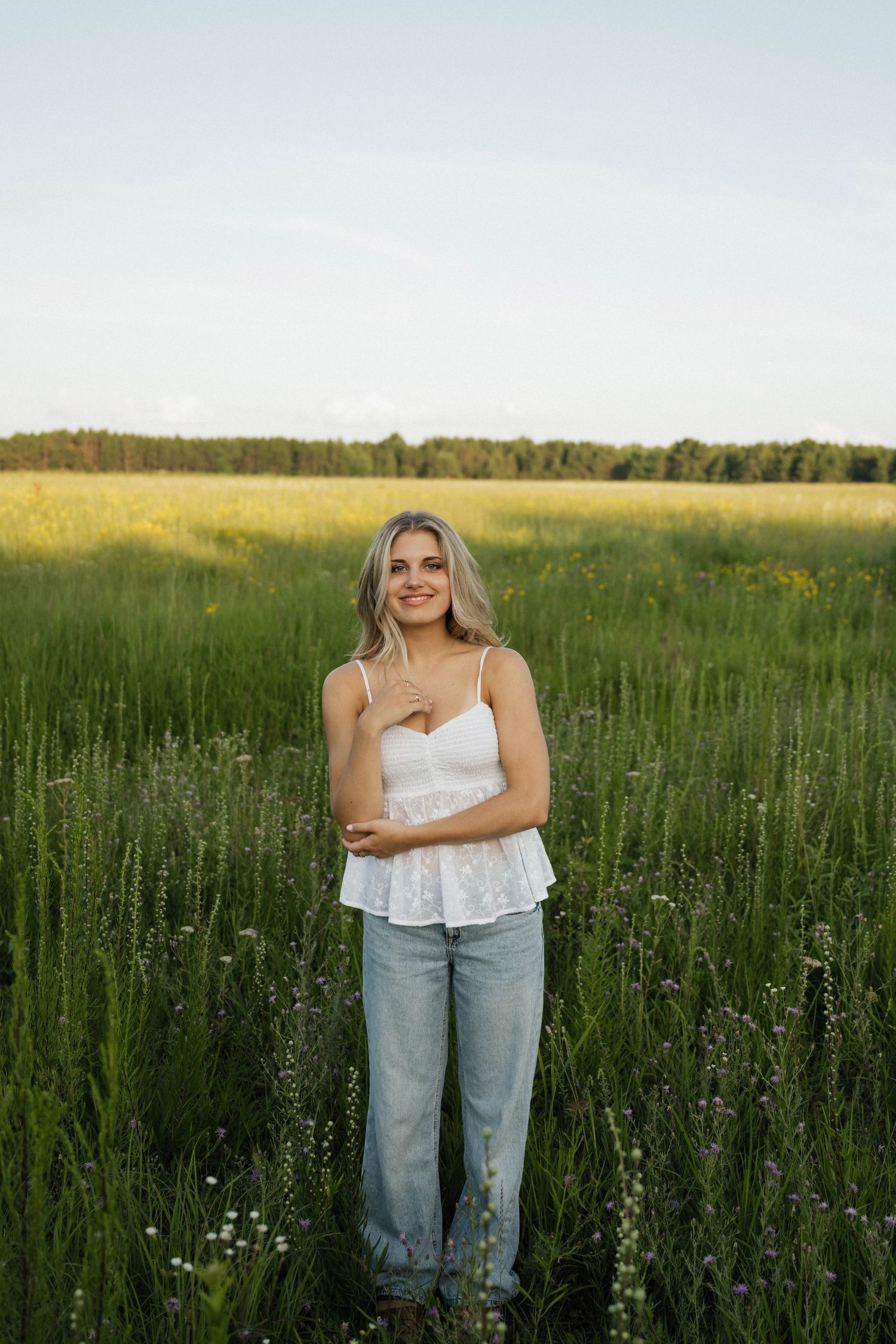 A smiling blonde woman in a white top and jeans stands in a vast green meadow under an open sky.