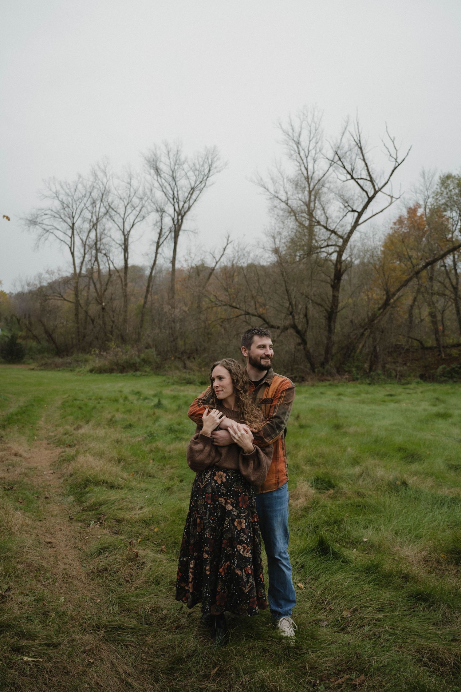 A couple embracing warmly in a green field surrounded by bare autumn trees on an overcast day.