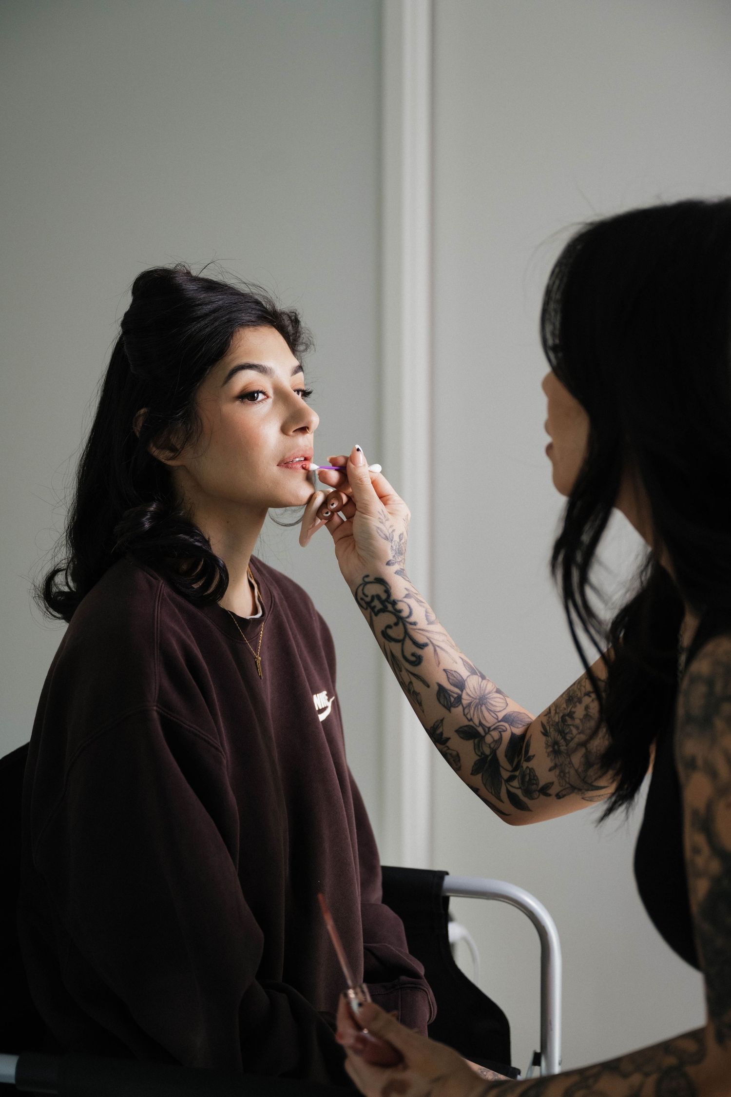 A tattooed makeup artist applies lipstick to a dark-haired bride during wedding day preparations.