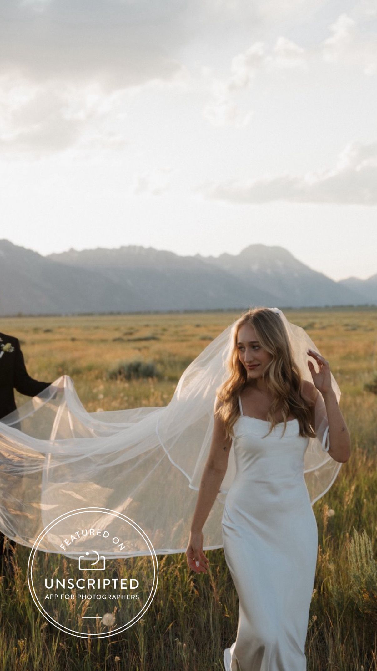 Elegant bride in white dress with long veil in scenic mountain field at sunset.