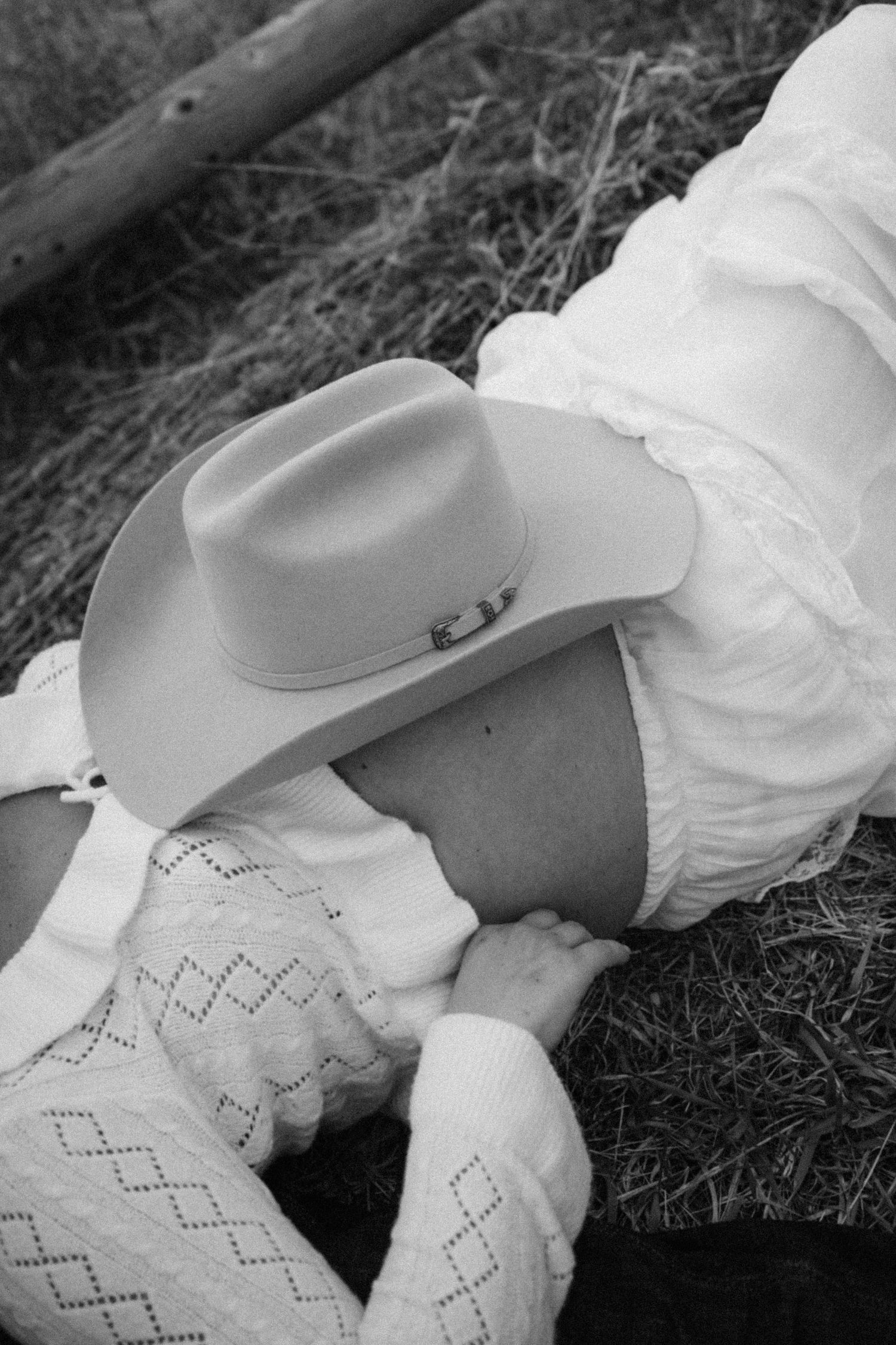 Black and white close-up of pregnant woman in cowboy hat and white lace dress lying in a field.