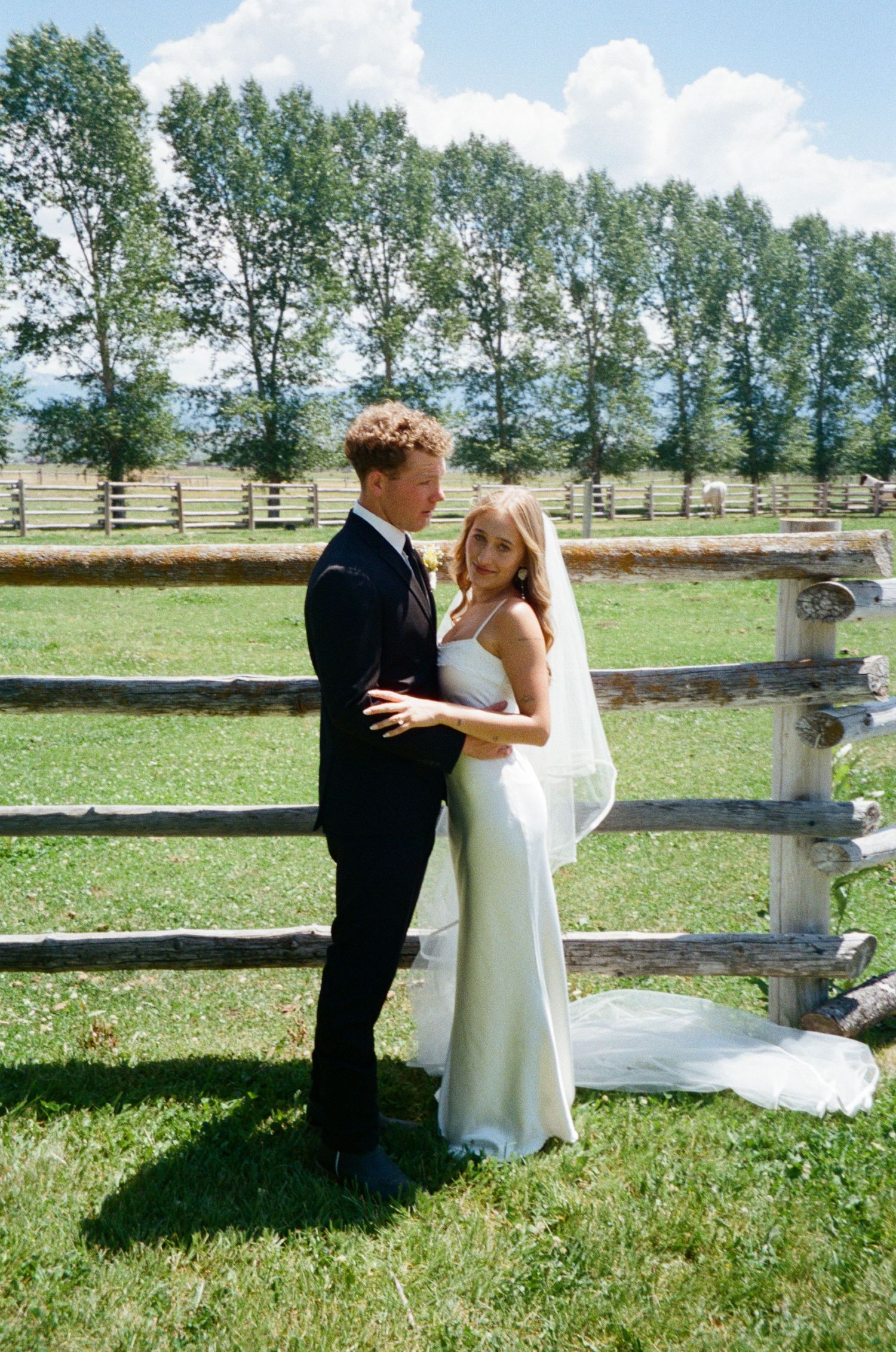 Bride in white slip gown with veil and groom in black suit embrace by rustic wooden fence on green lawn.