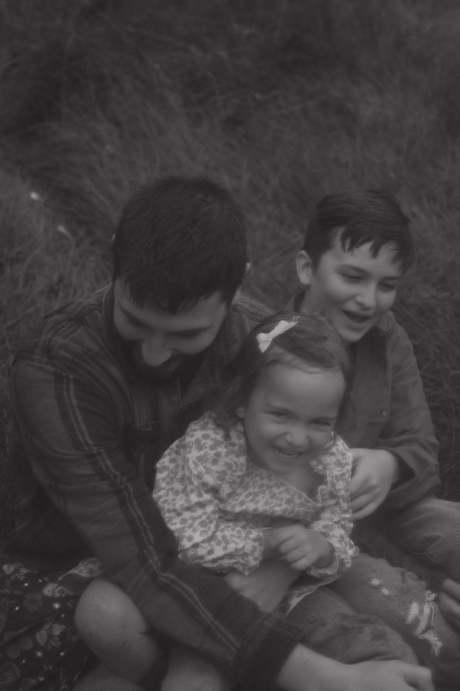 Black and white photo of a father laughing with his two young children sitting in the grass.