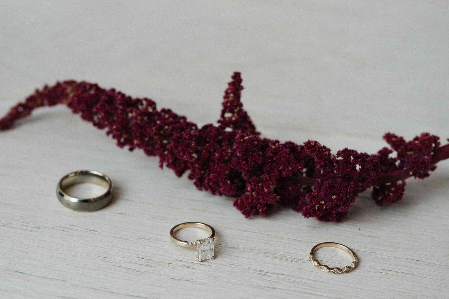 Wedding rings and a burgundy floral accent arranged on a white wooden surface.