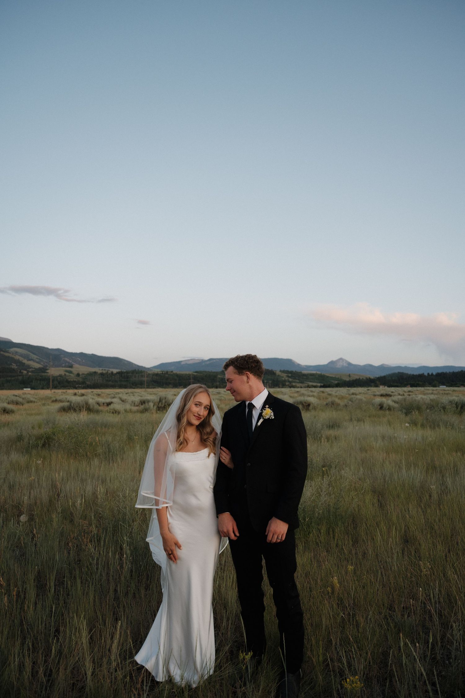 Bride and groom stand together in tall grass field with mountain range at dusk.