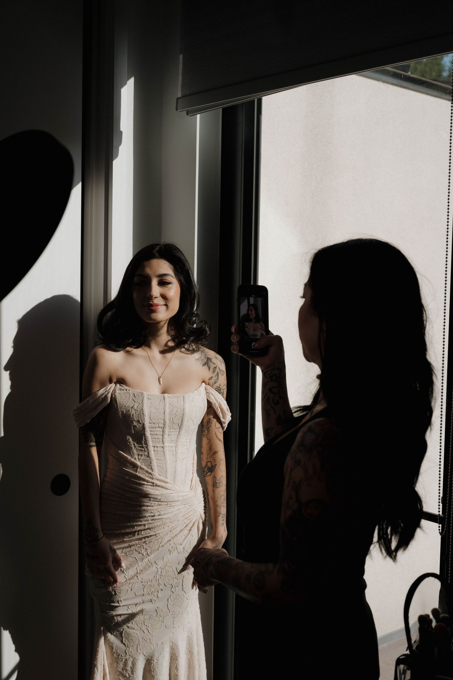 Bride in off-shoulder white gown being photographed by a silhouetted woman near a sunlit window.