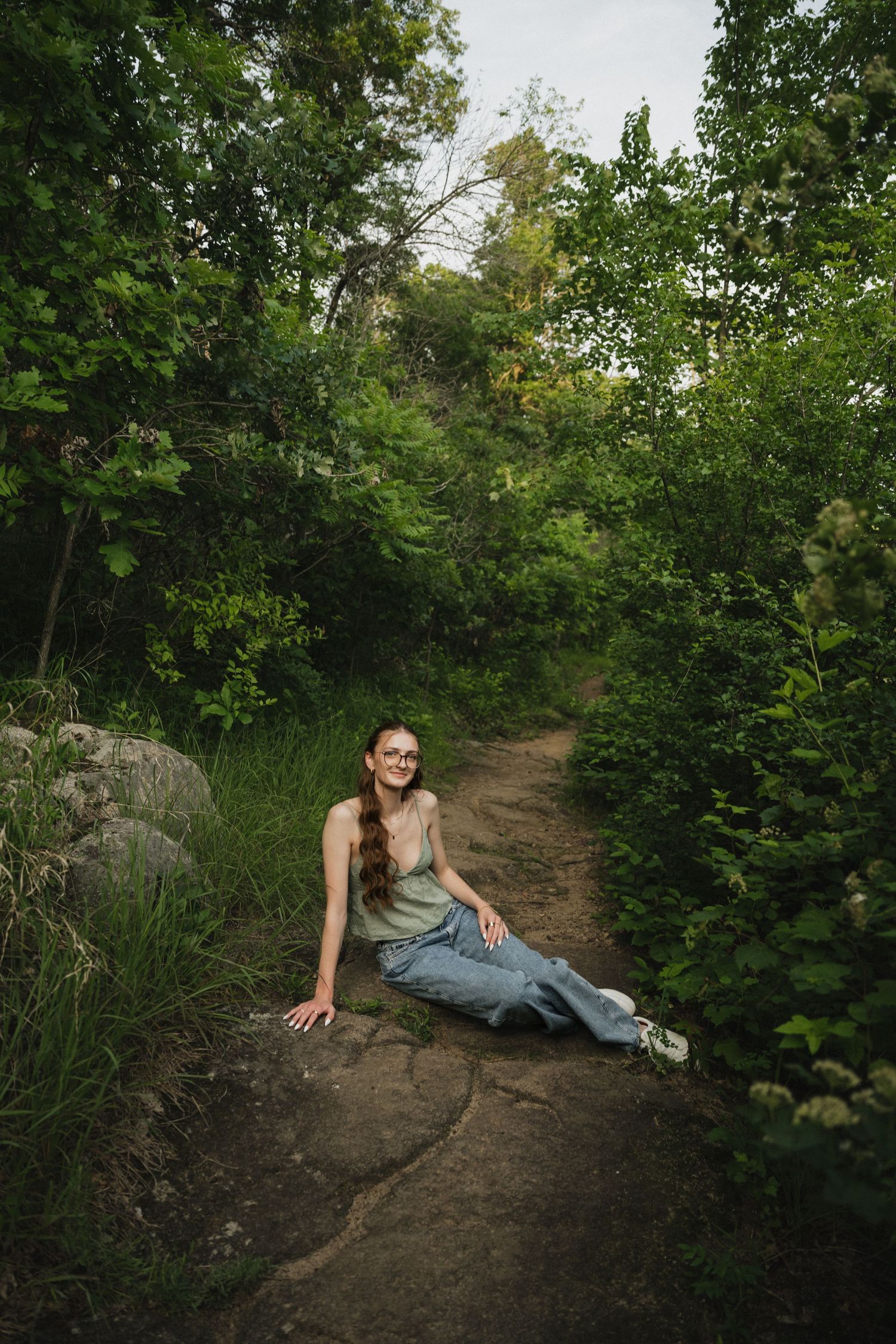 Smiling young woman with glasses sits on a rocky forest path surrounded by lush green trees and vegetation.