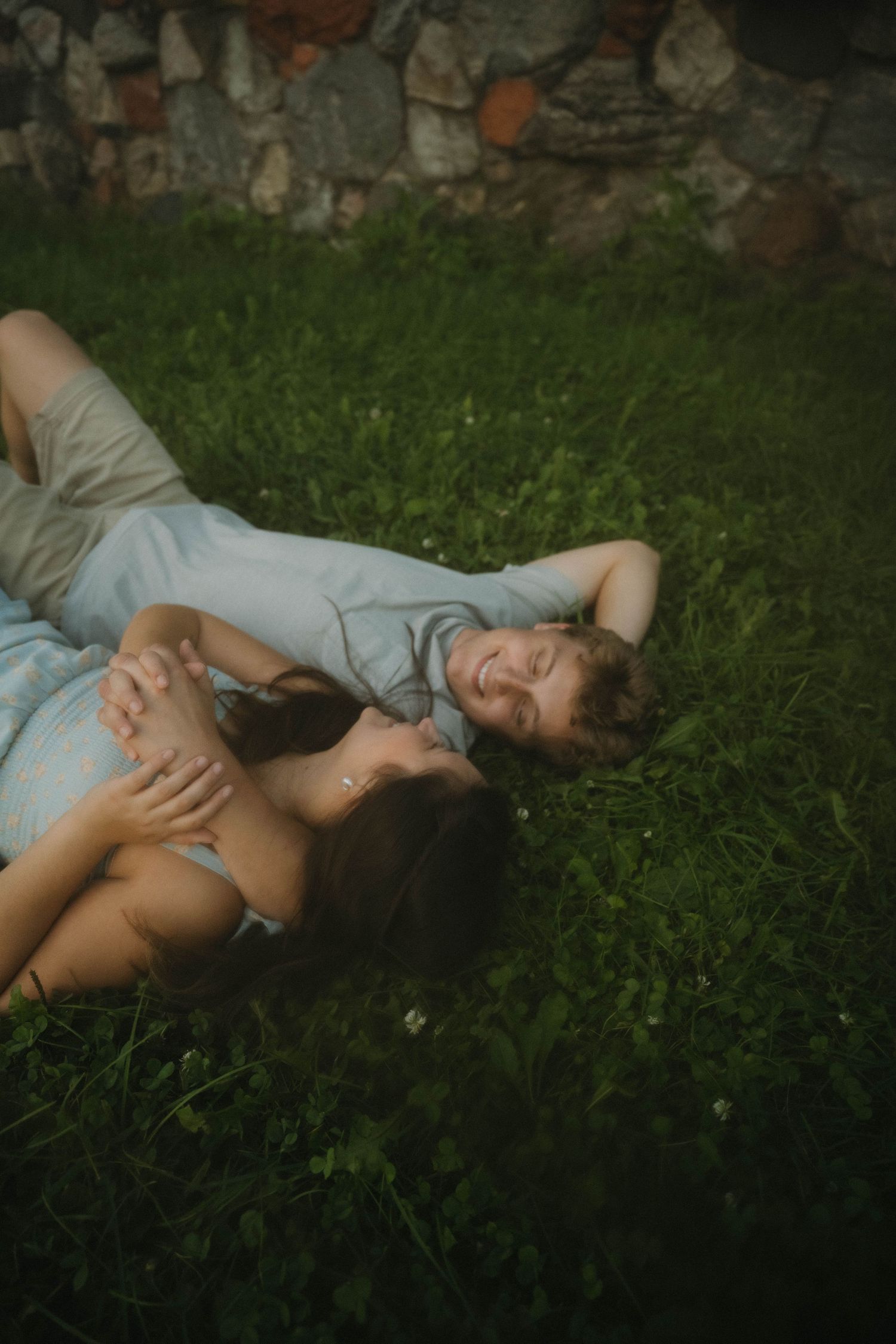 Couple lying on green grass laughing together near stone wall in soft natural light.