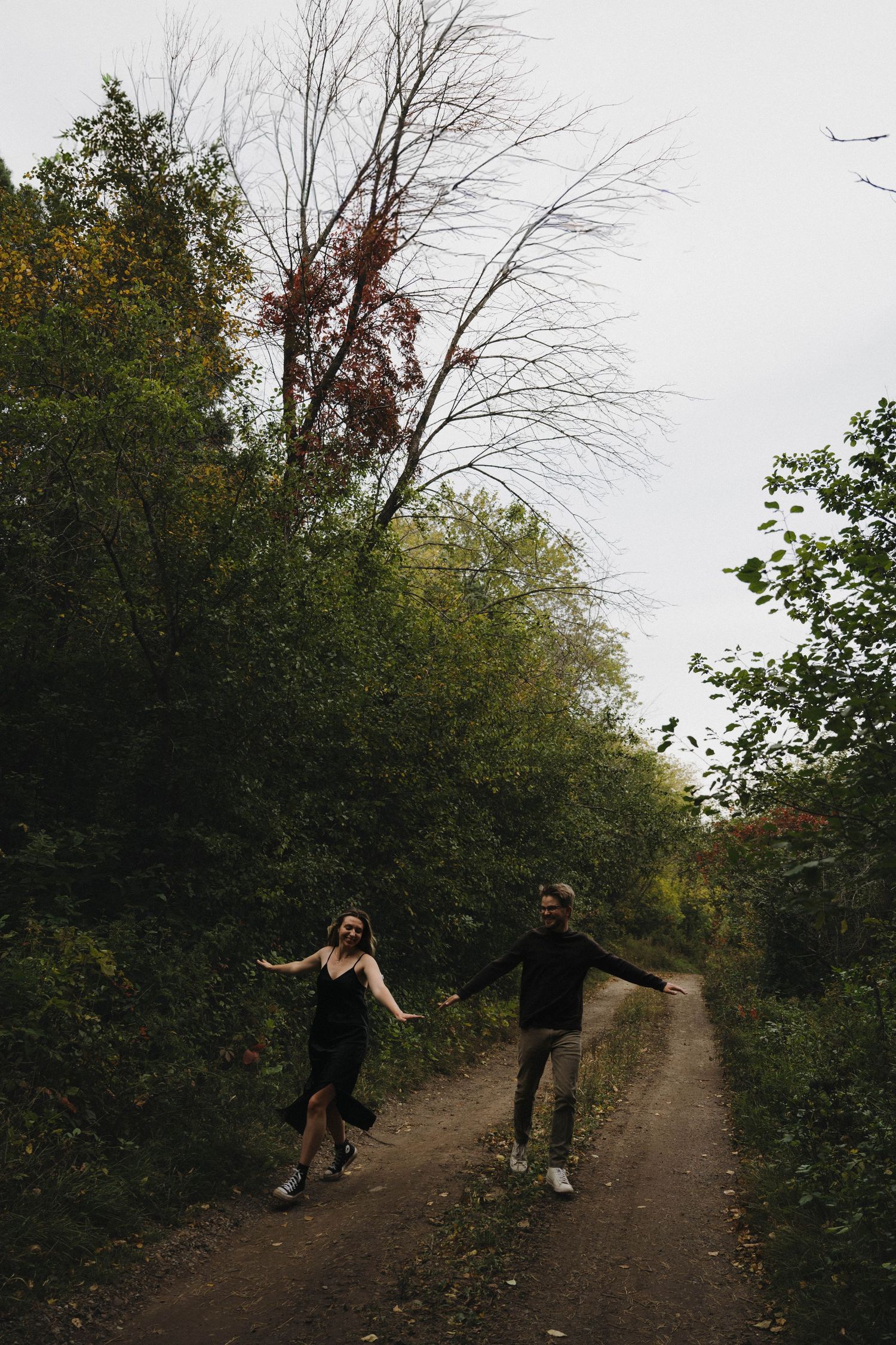 A joyful couple dancing and spinning on a rustic dirt path surrounded by autumn trees.