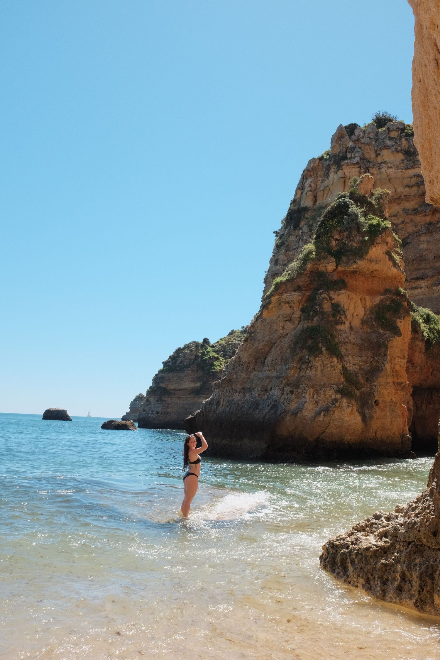 Woman in black bikini standing in turquoise water beside towering orange cliffs at Algarve beach Portugal.
