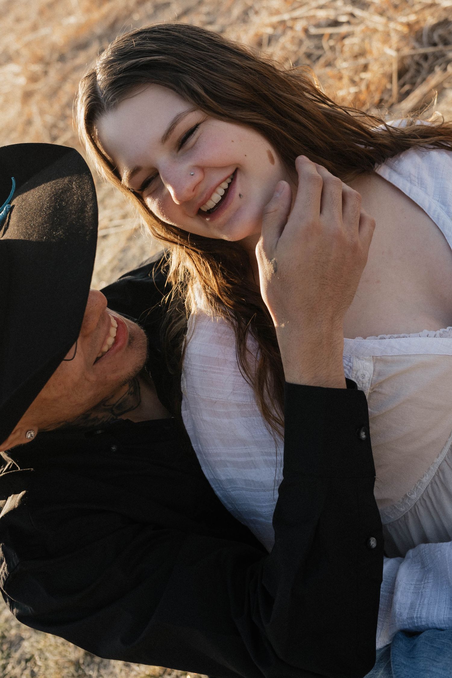 Joyful young woman laughing as a man in a black hat embraces her in a golden sunlit field.