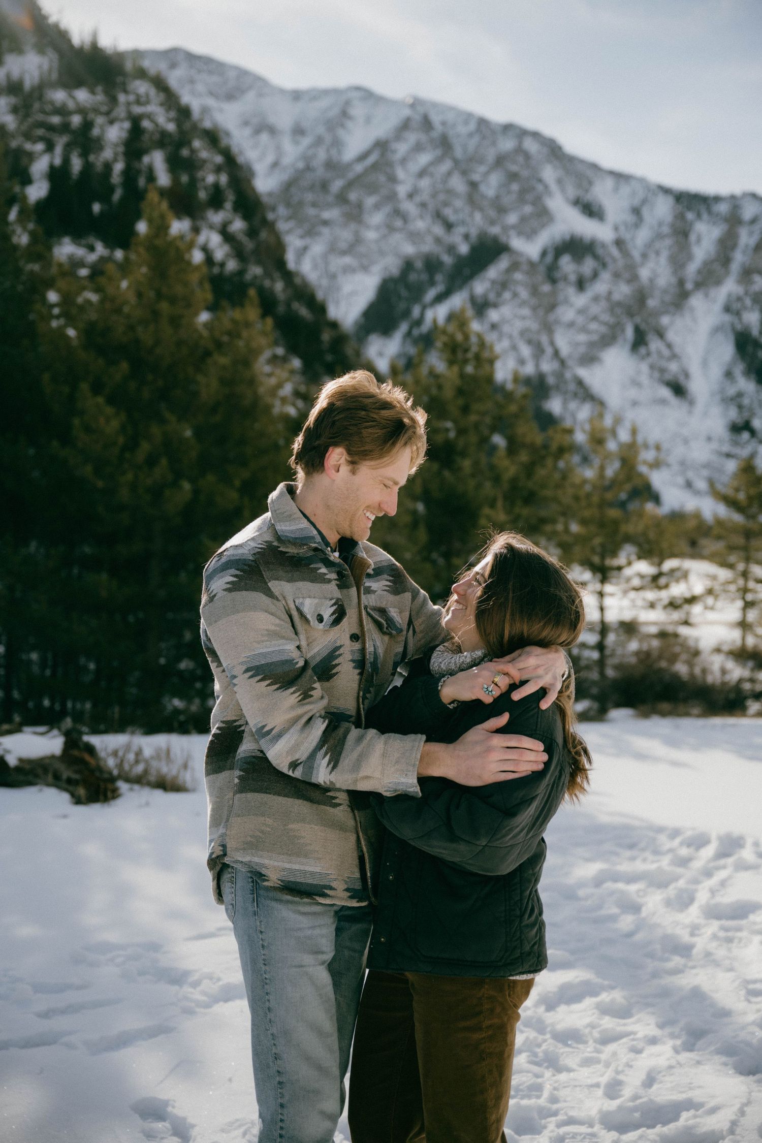 Happy couple embraces and laughs together in sunny snowy mountain setting with pine trees behind them.