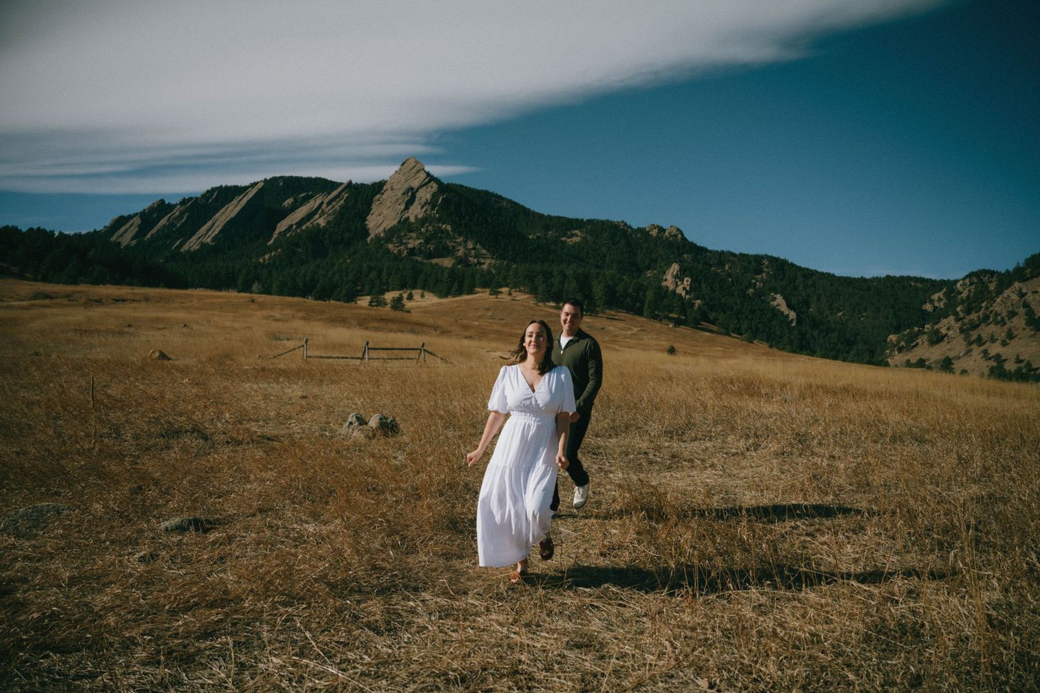 Couple walks hand in hand through golden meadow with stunning Colorado Flatirons mountains behind them.
