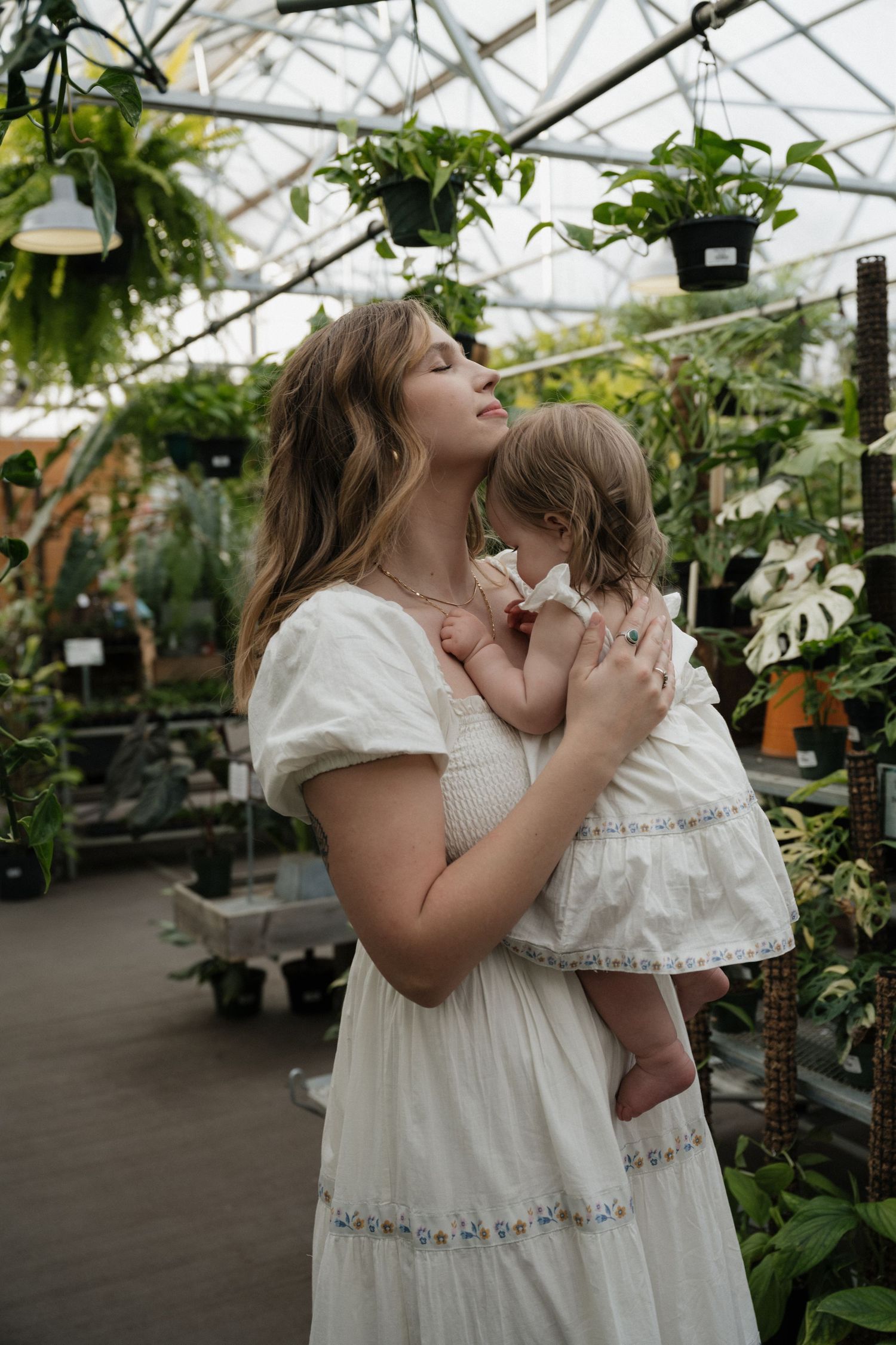 A mother in a white dress tenderly holds her young daughter in a lush greenhouse filled with tropical plants.
