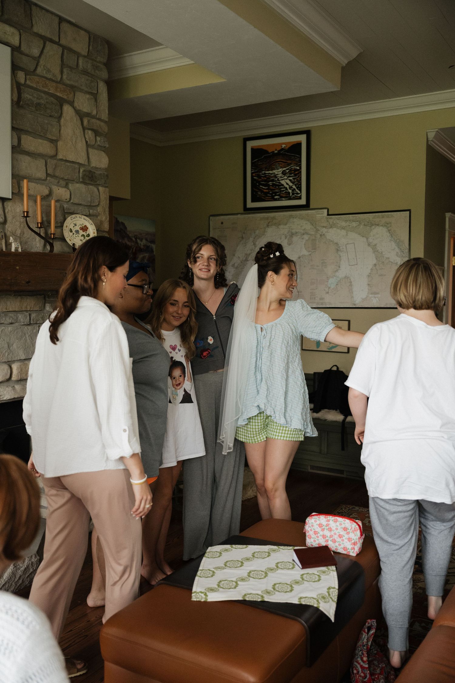Bride in veil surrounded by smiling bridesmaids and family in a cozy stone fireplace room.