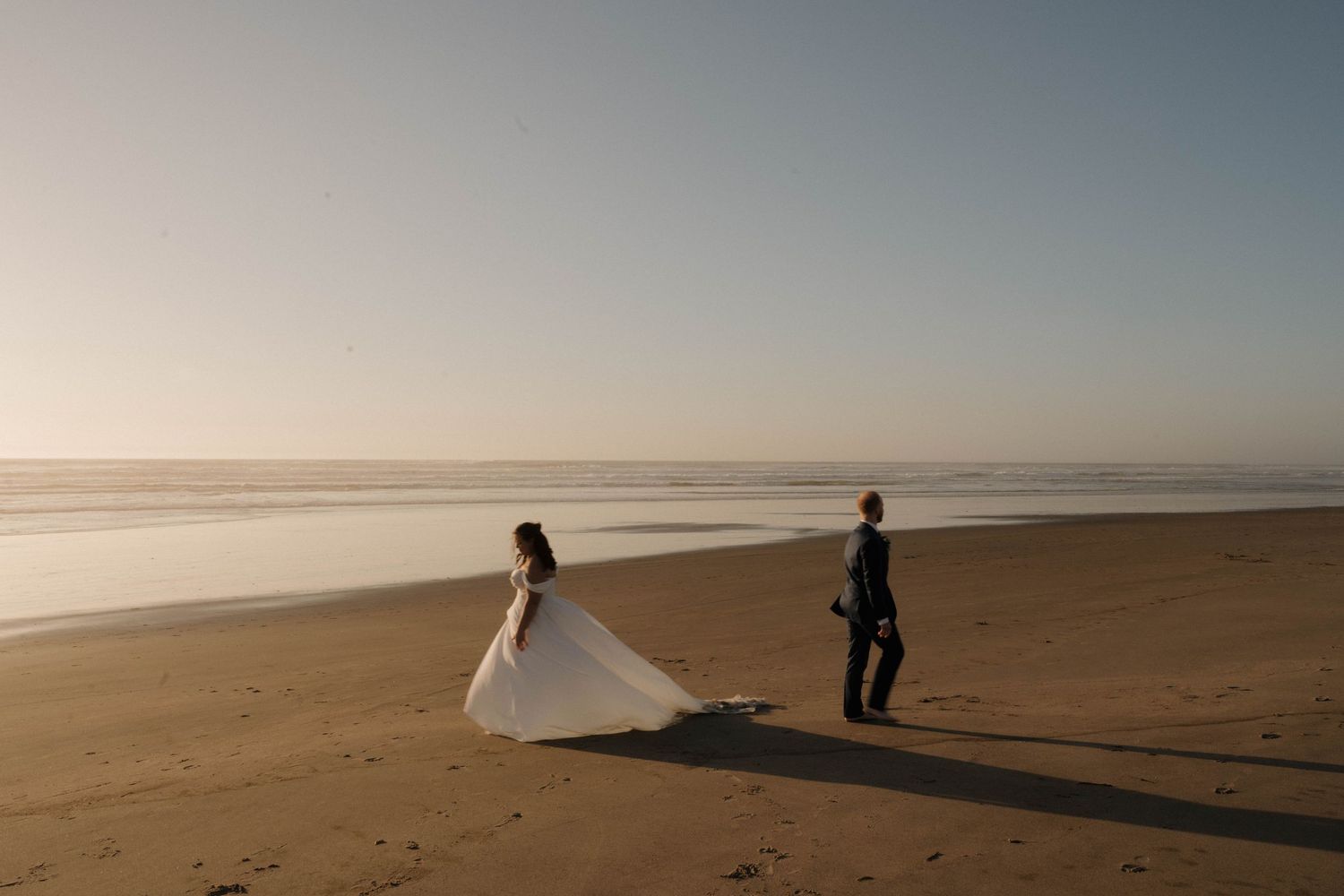 Bride in elegant white gown and groom walk along a serene sandy beach at golden sunset hour.