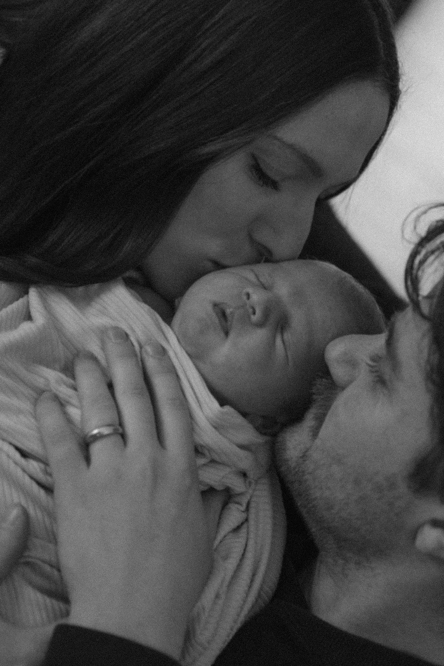 Black and white photo of parents kissing their swaddled newborn baby together.
