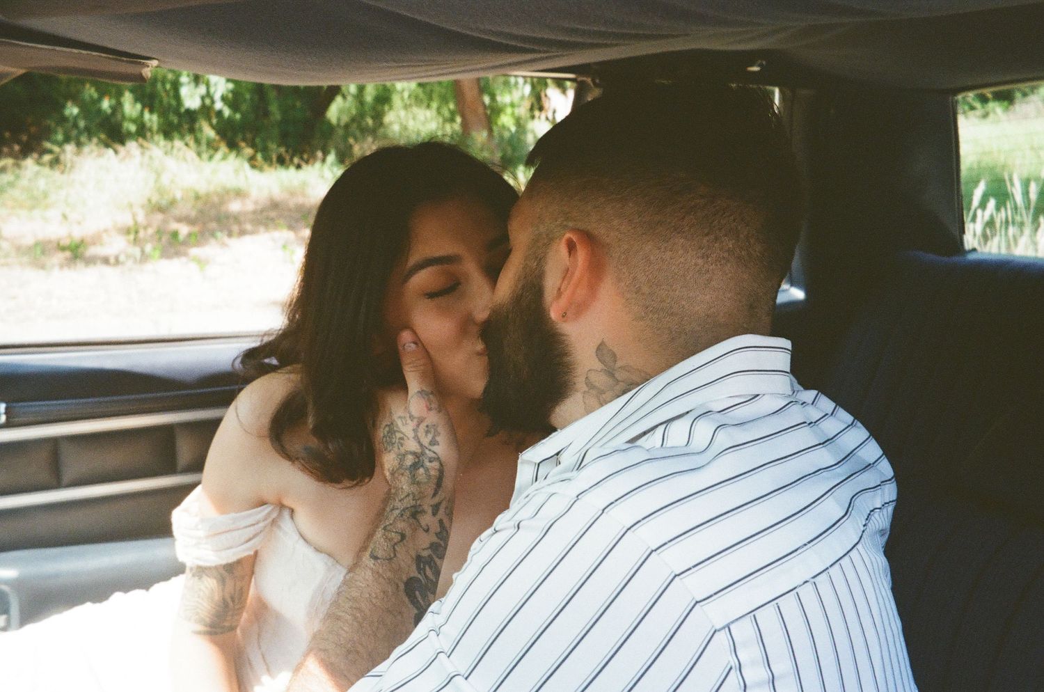 Couple shares a kiss inside a vehicle framed by green trees in a romantic film-style photo.