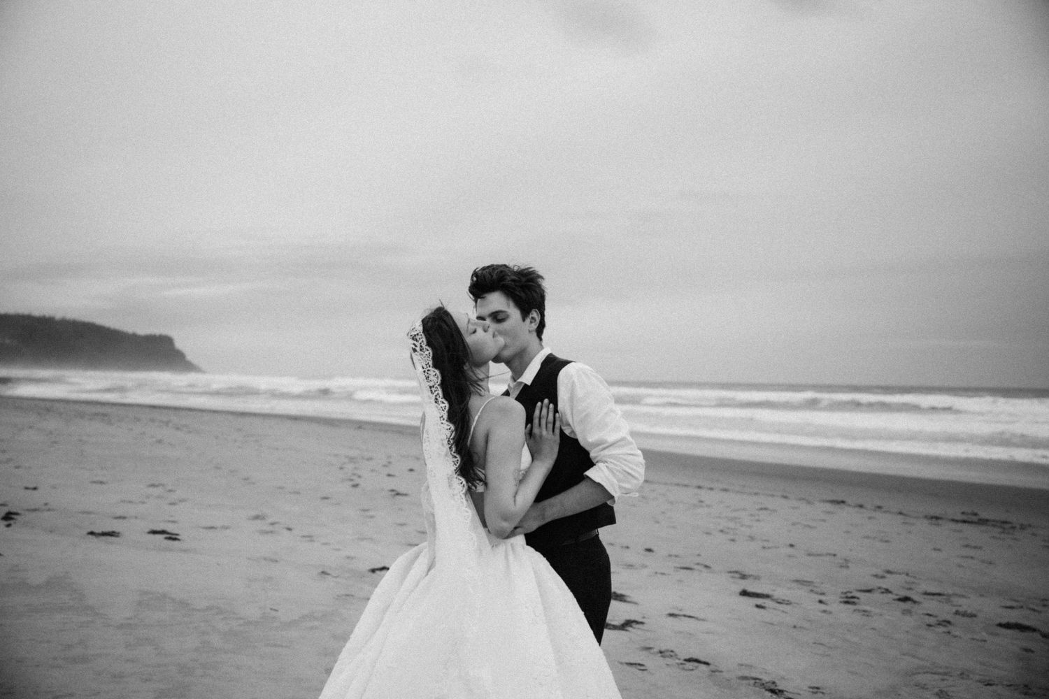 Black and white romantic beach wedding photo of bride and groom kissing by ocean waves.